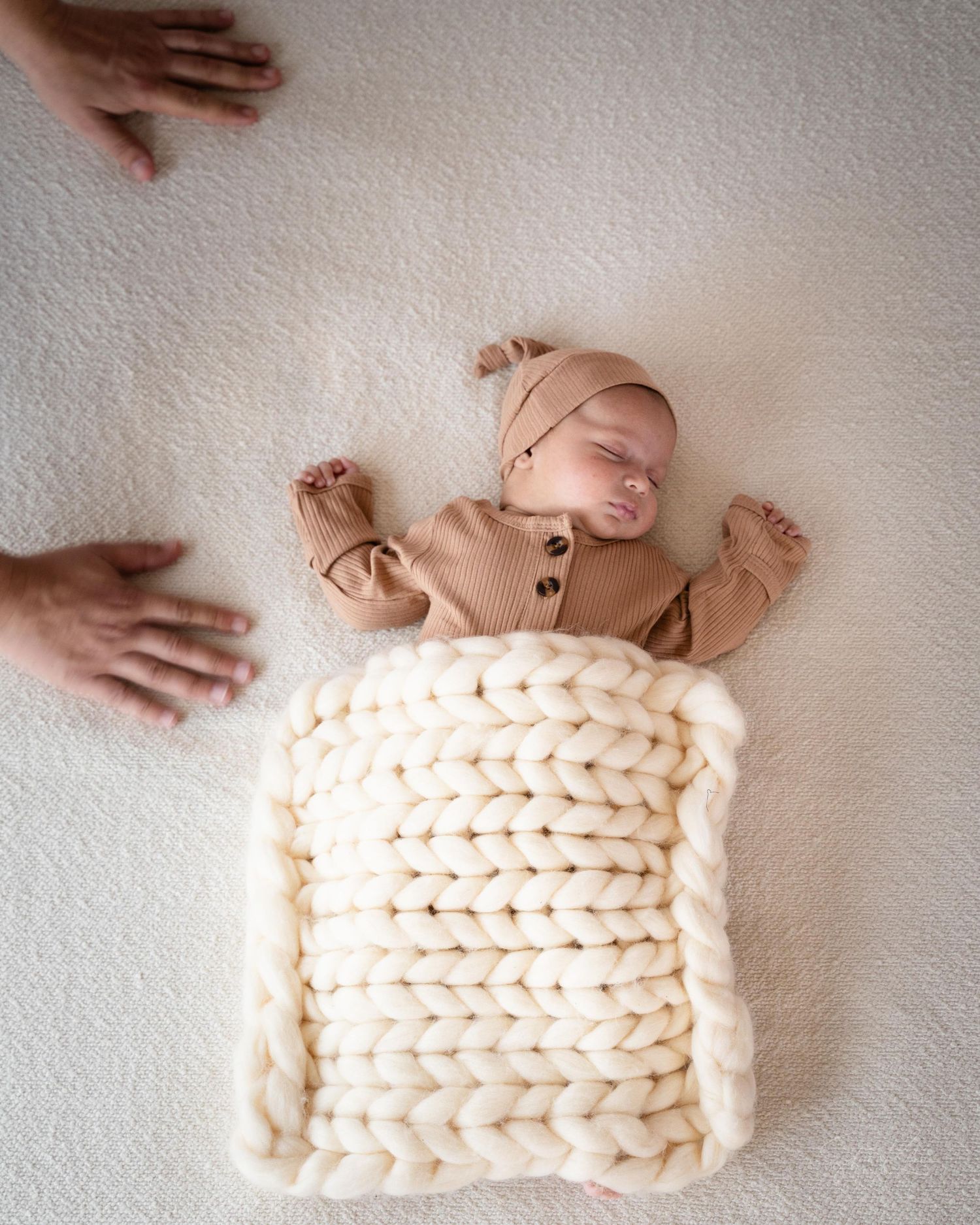 A sleeping newborn on chunky knit cream blanket with adult hands reaching in from sides of frame.