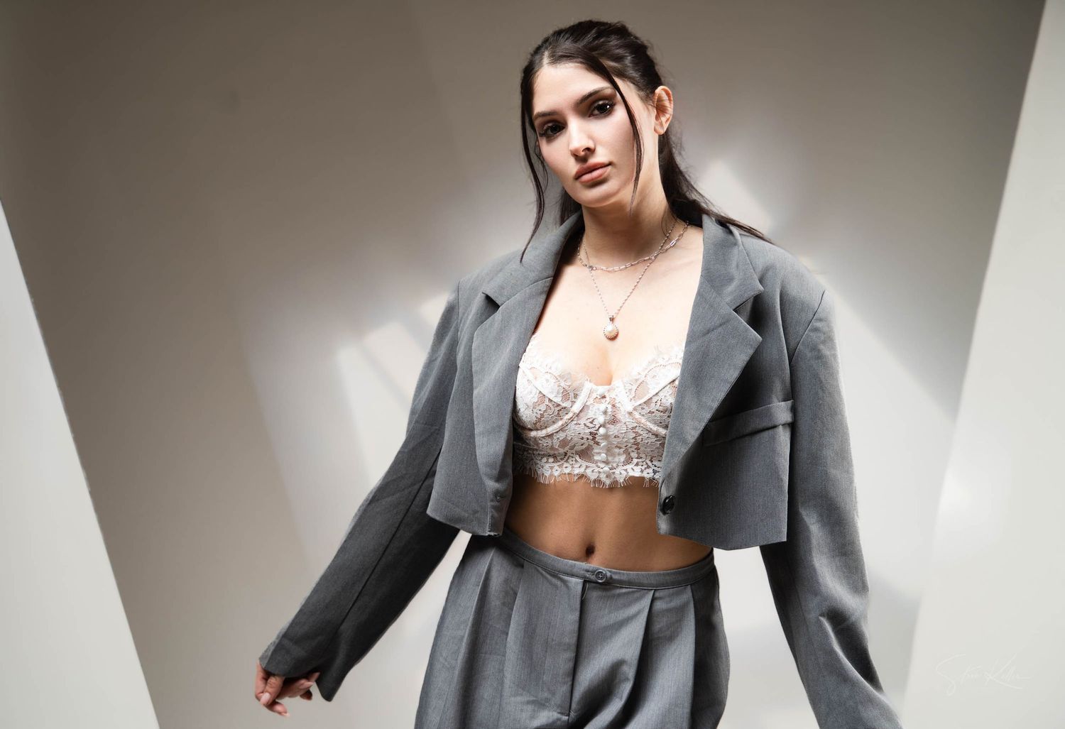 Fashion model wearing a cropped gray blazer with white lace bralette against a light background.