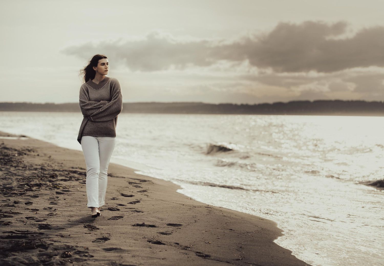 Person in casual attire takes a solitary stroll along a sandy beach with moody overcast skies.