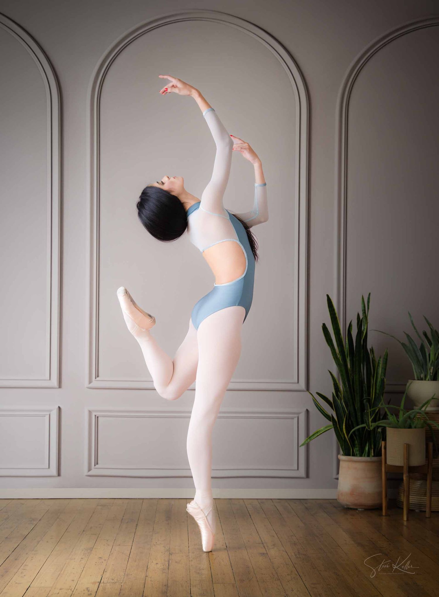 Ballet dancer in light blue leotard performs graceful pose en pointe against white paneled wall with potted plant.