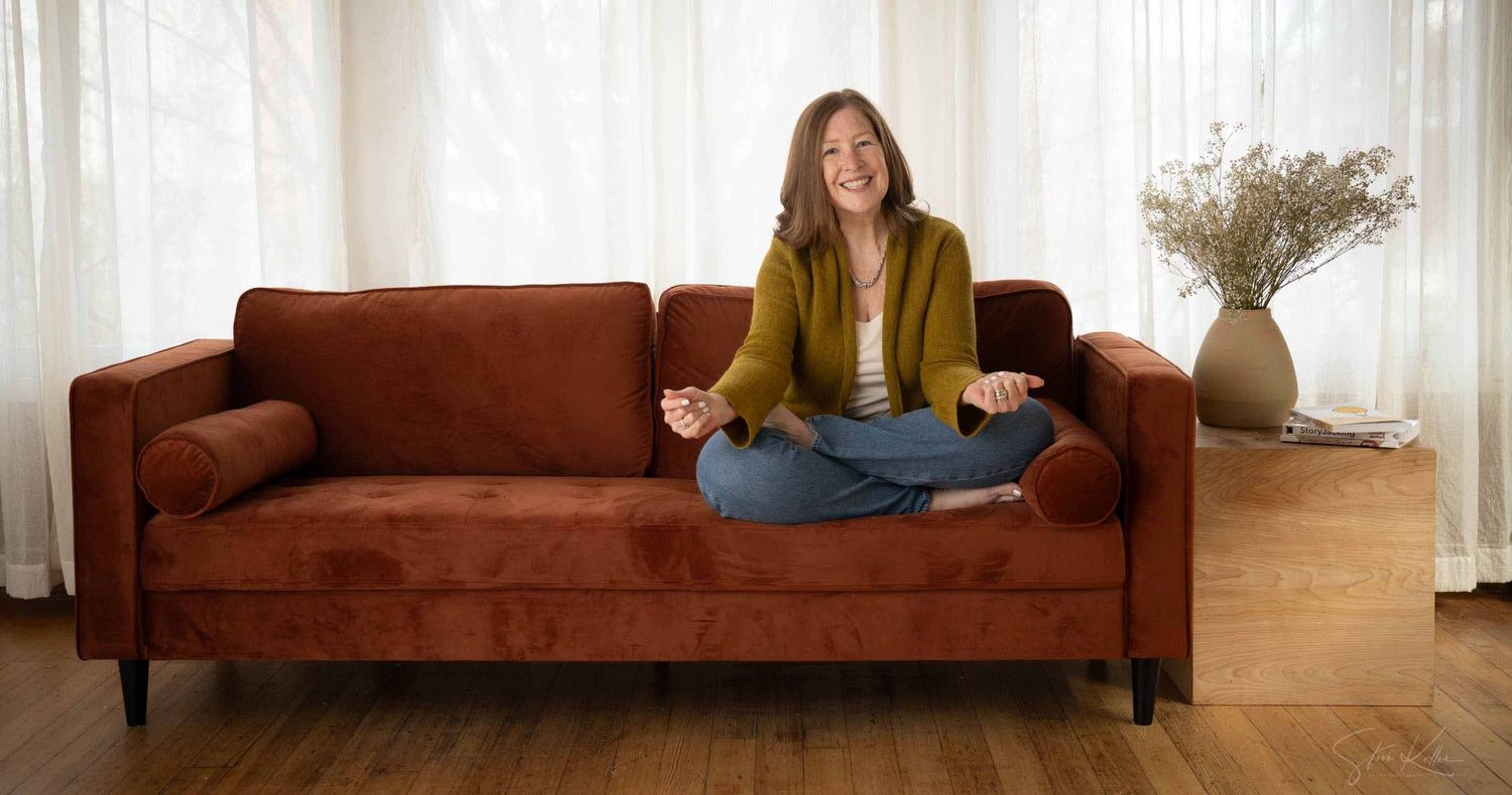 Smiling woman in a green cardigan sits cross-legged on a rust velvet sofa beside a vase of dried flowers in a bright, airy living room.
