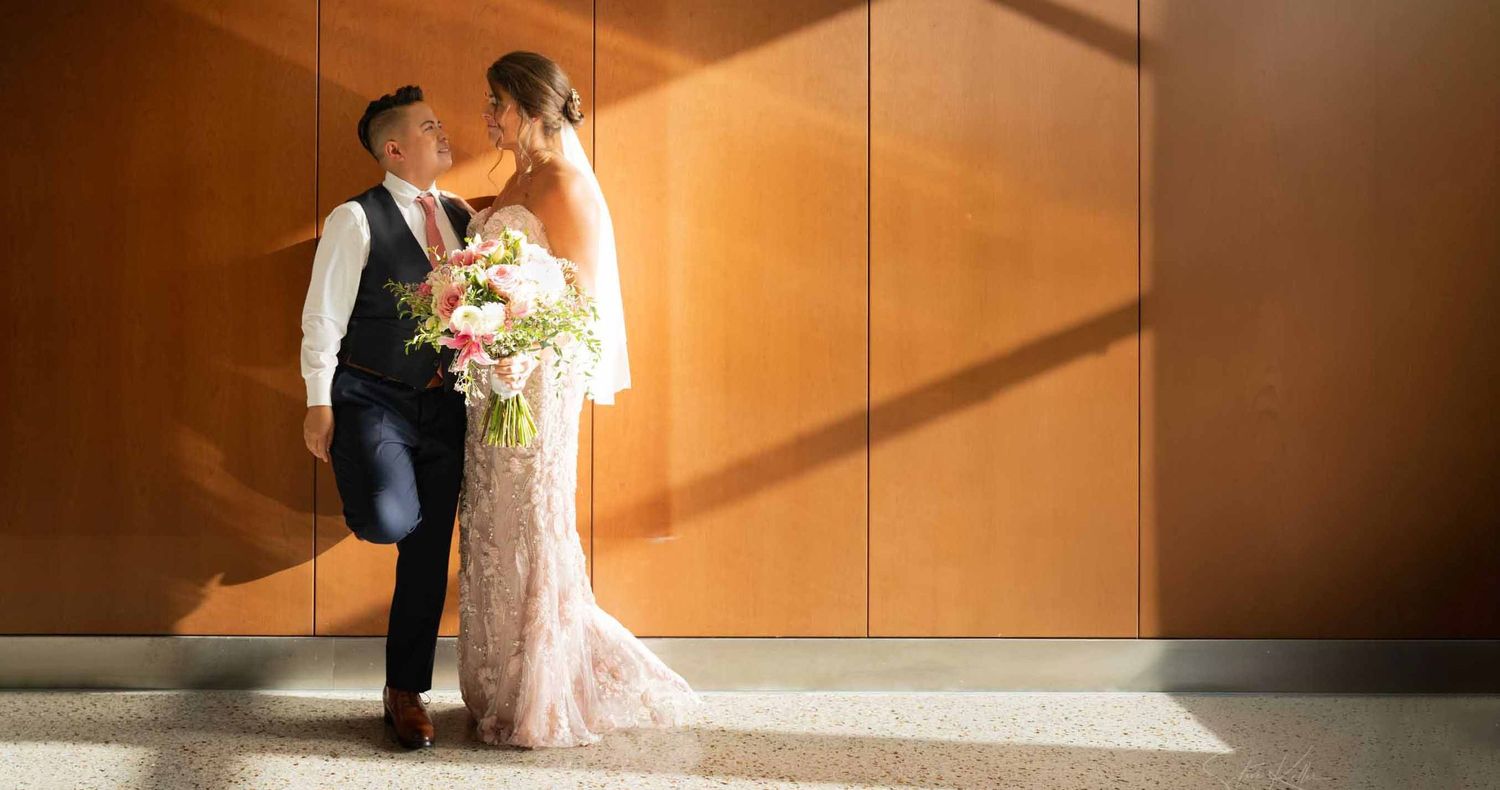 Bride in a lace wedding gown holding a pink bouquet leans on her groom in a vest as warm sunlight streams across a wood-paneled wall.