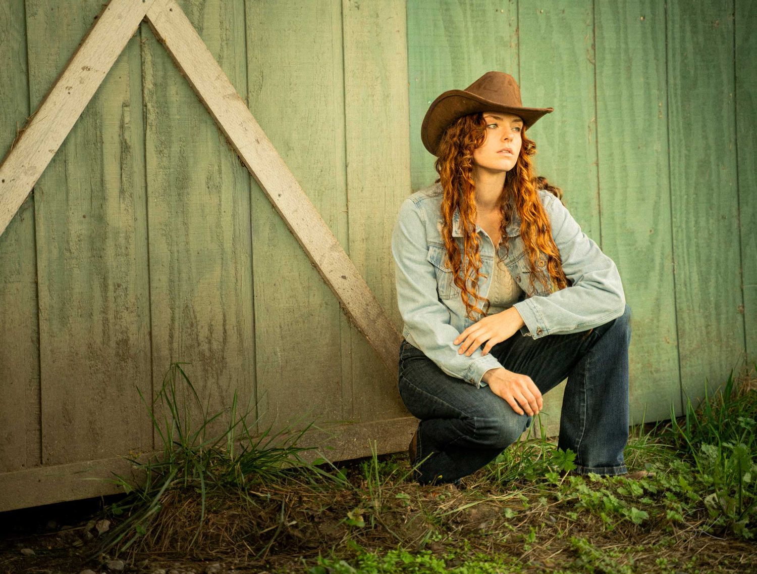 Woman in cowboy hat with long curly red hair sits against rustic barn wall in jeans and denim shirt.