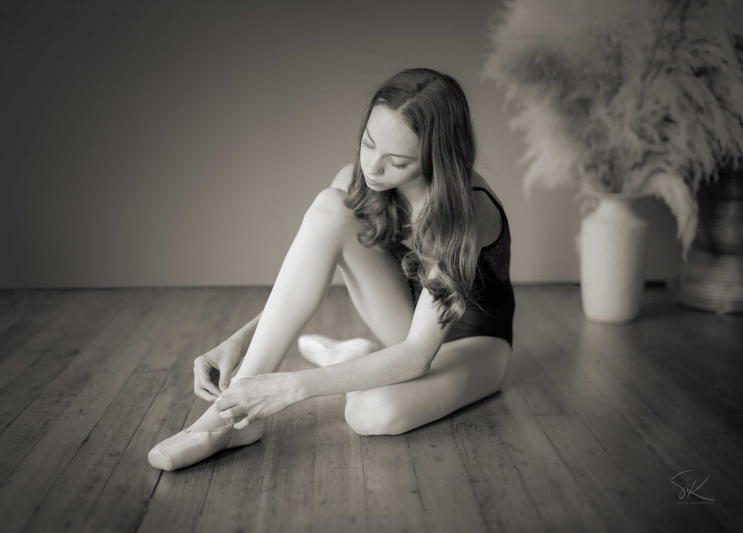 Black and white photo of a ballerina sitting on wooden floor tying her pointe shoes in a studio.
