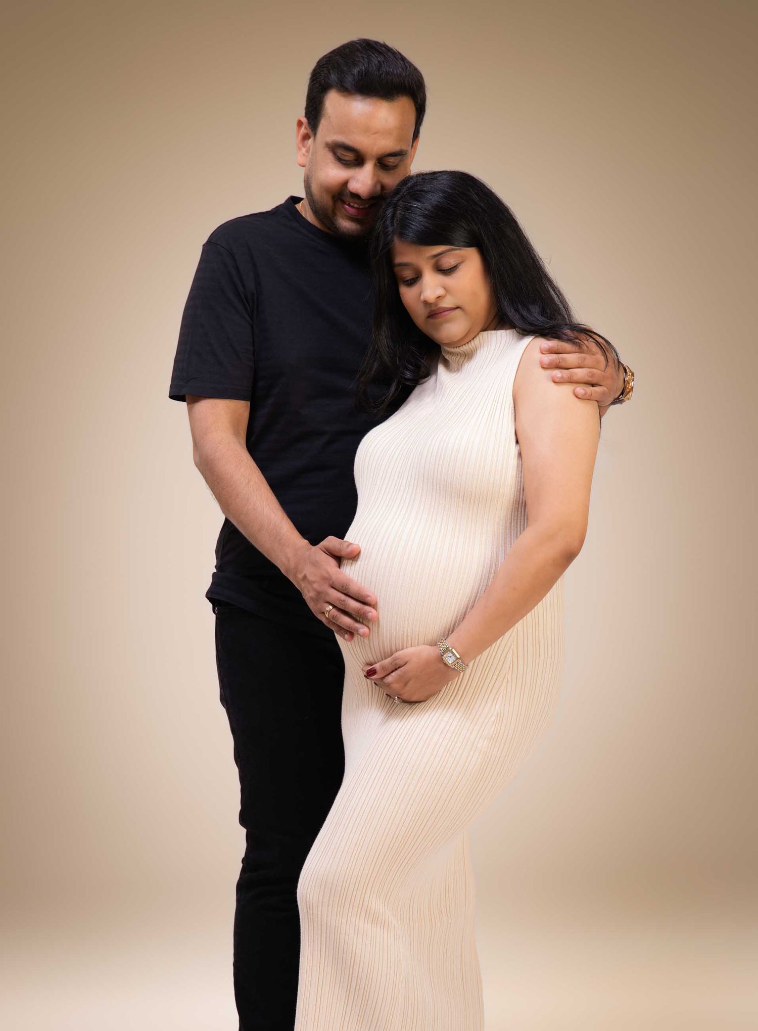 Couple in maternity photoshoot, man in black embracing pregnant woman in white ribbed dress, studio portrait.