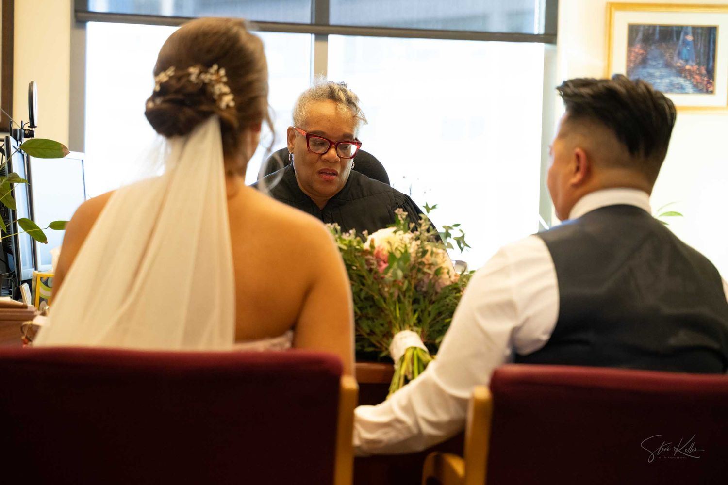 A bride and groom sit at a table with flowers while an officiant speaks during an intimate indoor wedding ceremony.