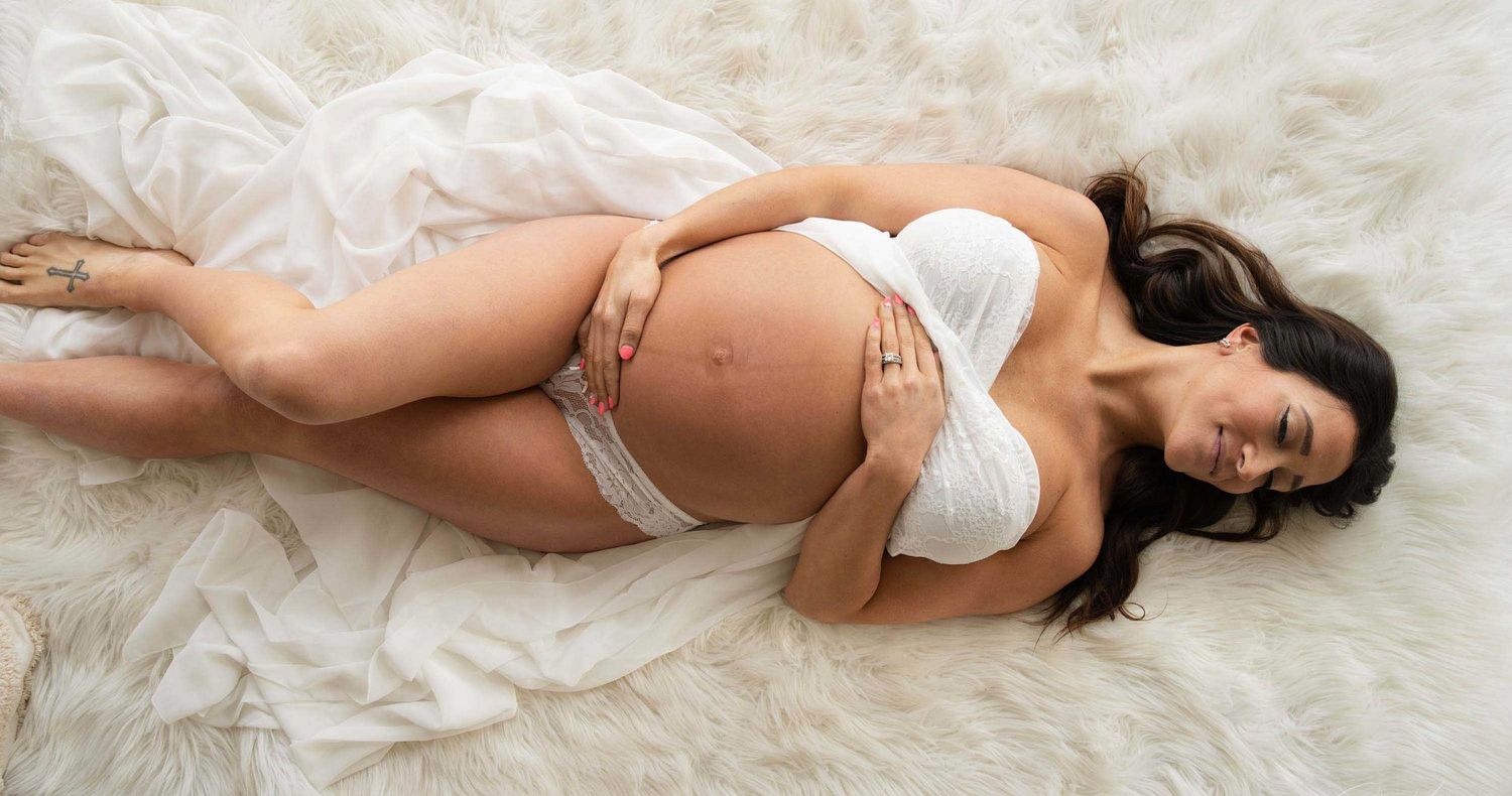 Pregnant woman in white lingerie lying on a soft fur rug, cradling her bare baby bump in a maternity photoshoot.