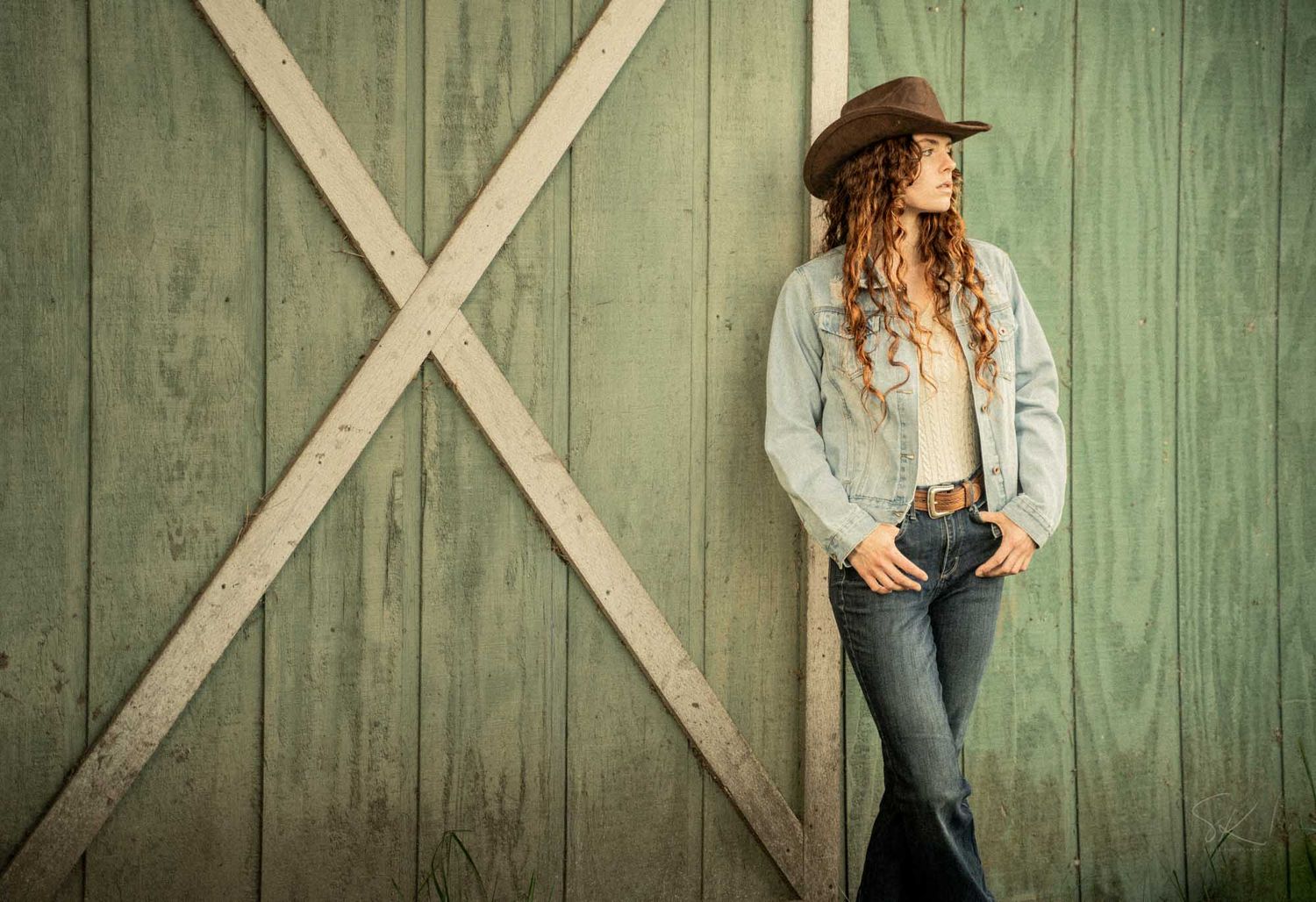 Young woman in cowboy hat, jeans, and denim jacket posing against a rustic green barn door.