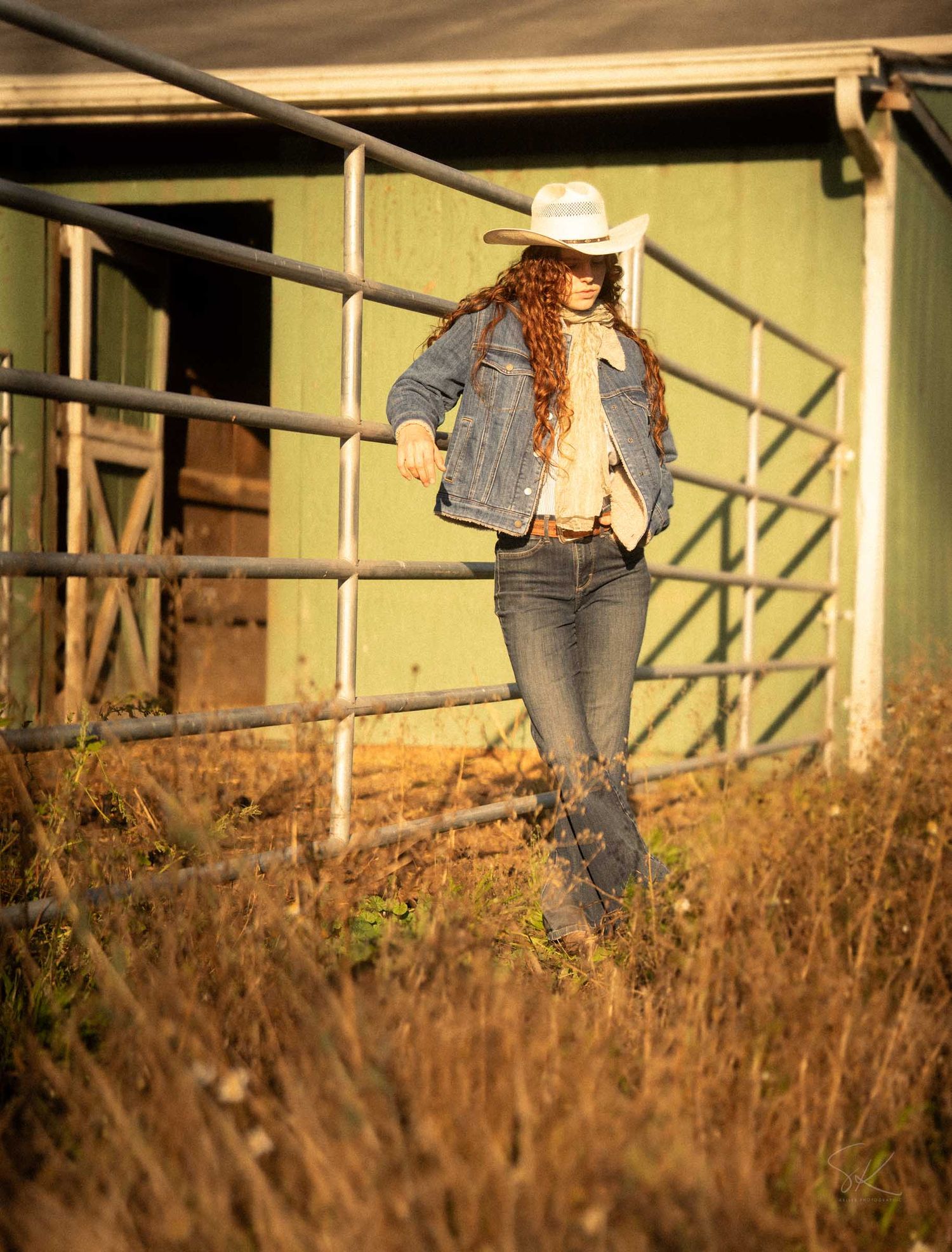 A woman in a denim jacket, jeans, and white cowboy hat walks near a metal gate by a green barn.