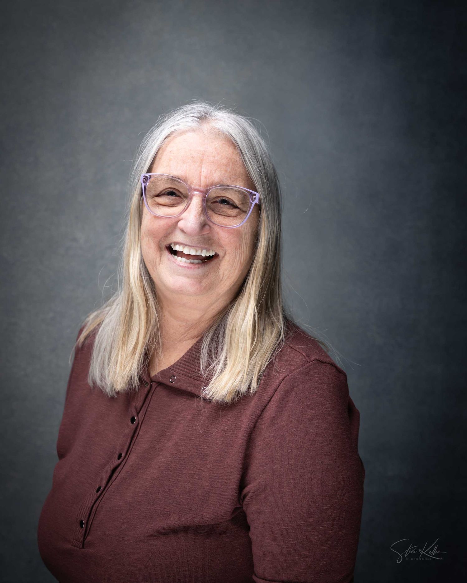 Smiling older woman with gray hair and purple glasses wearing a burgundy top against a gray studio background.