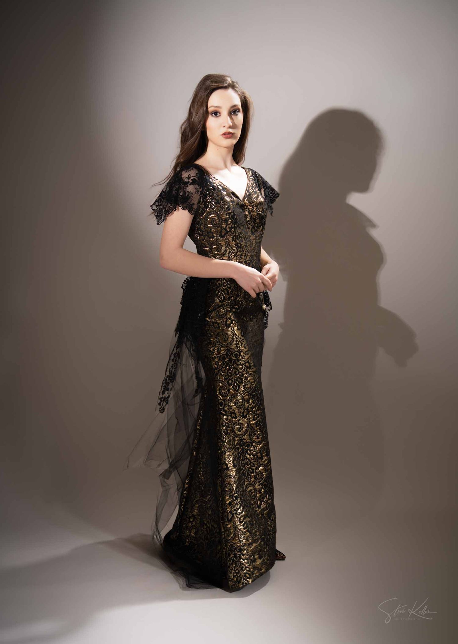 Elegant black and gold lace evening gown with cap sleeves and dramatic silhouette against grey studio backdrop.