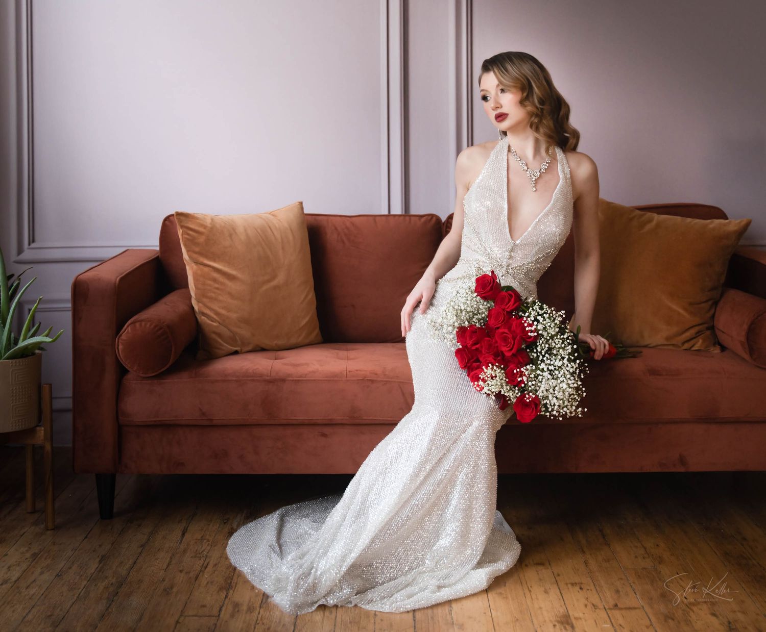 A bride in a vintage-style lace wedding gown sits elegantly on a rust velvet couch holding a vibrant red rose bouquet.