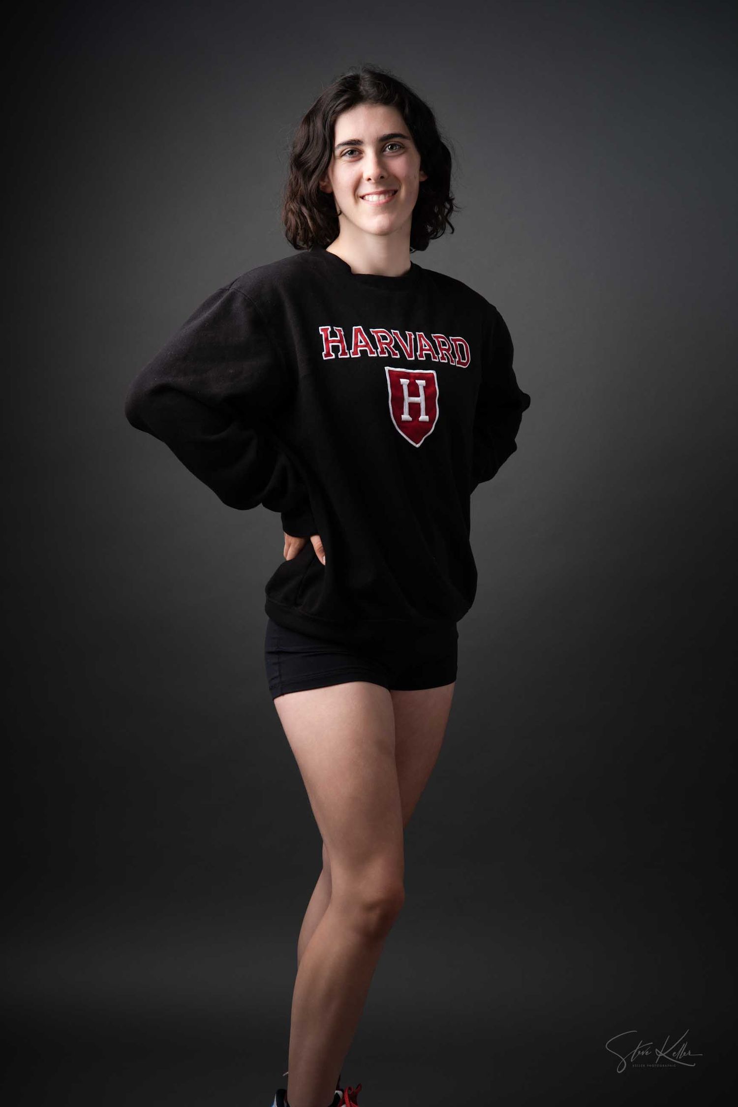 A person poses in a black Harvard University sweatshirt and shorts against a dark studio backdrop.