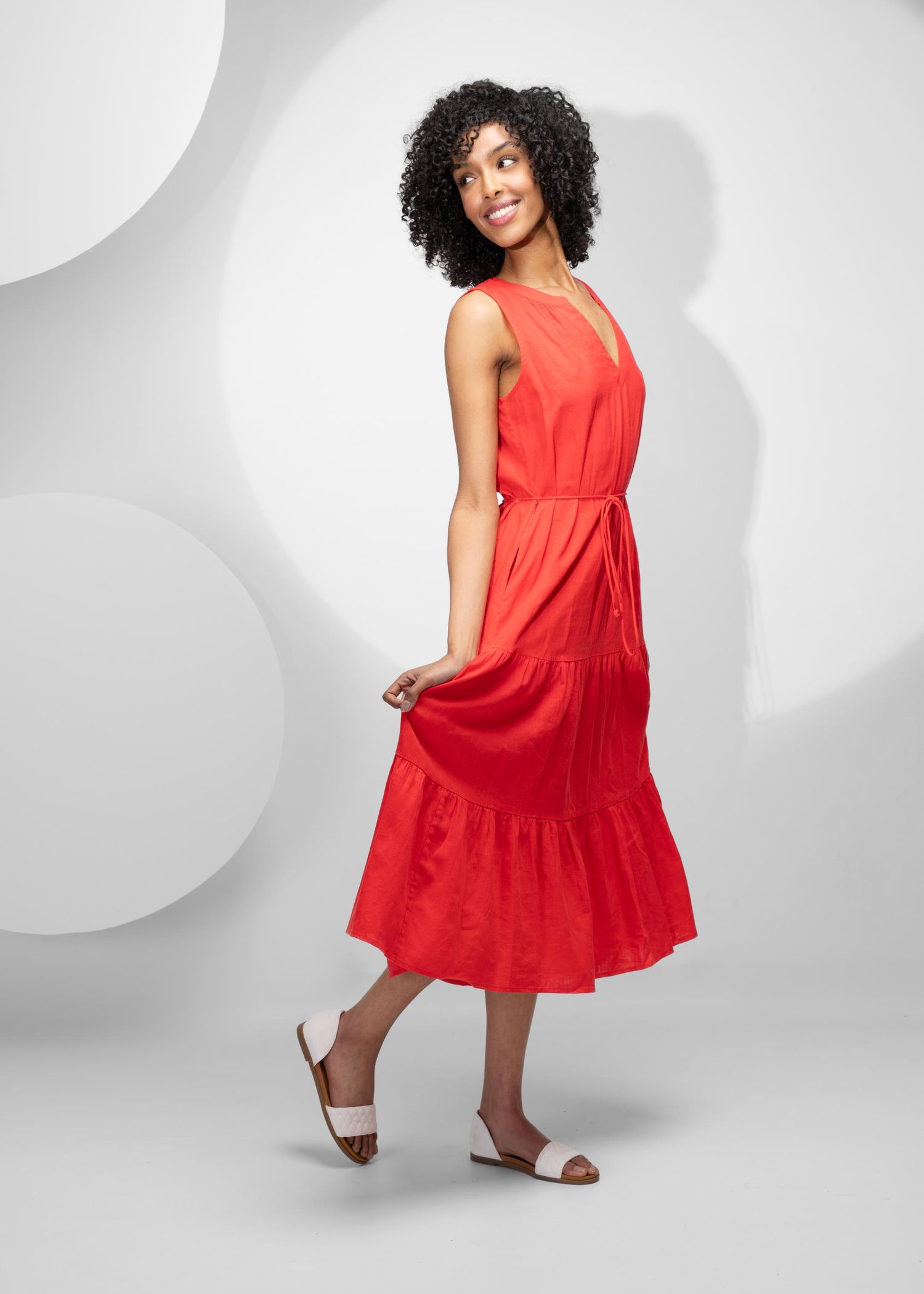 A sleeveless red midi dress with tiered skirt and brown sandals photographed against a gray background with circular patterns.