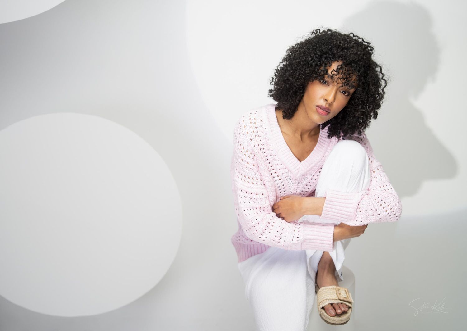 Woman with curly hair wearing pink cardigan and white dress poses against white backdrop.