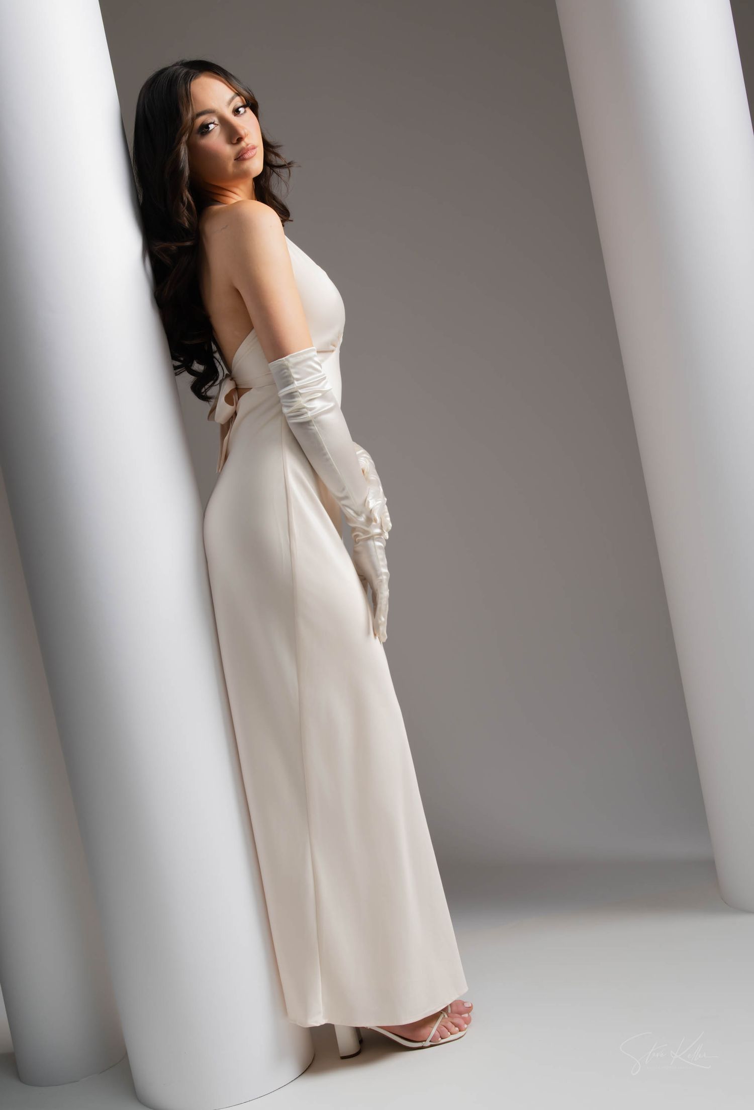 Elegant white formal gown with long gloves photographed against a minimalist grey and white backdrop in a studio setting.