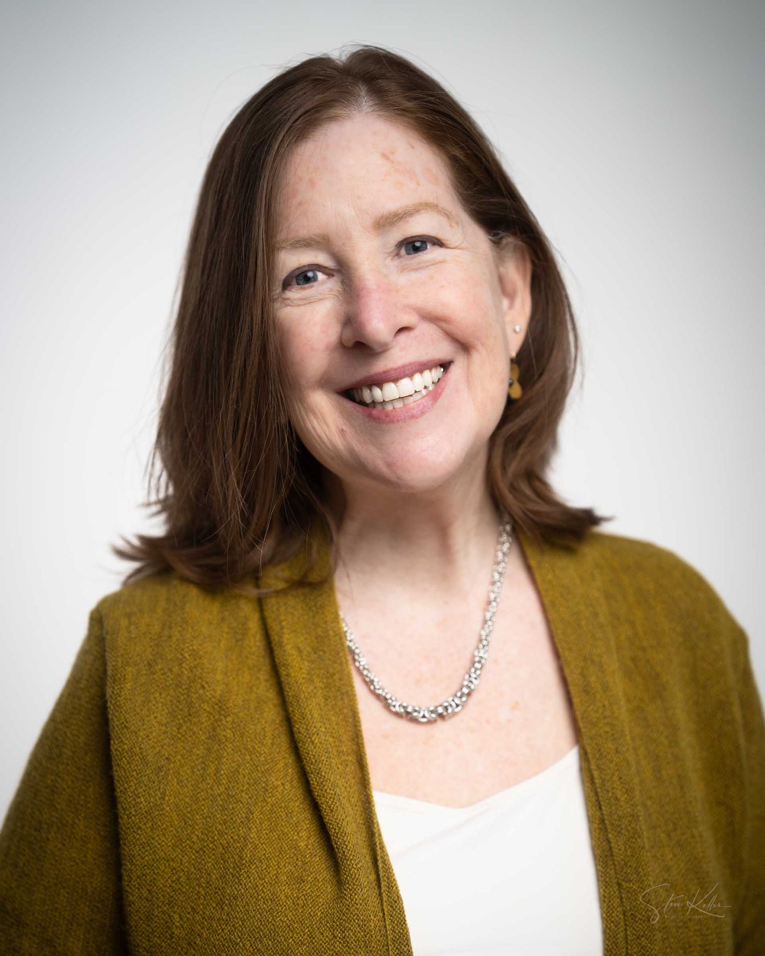 Professional headshot of a smiling woman with brown hair wearing a mustard cardigan and pearl necklace.