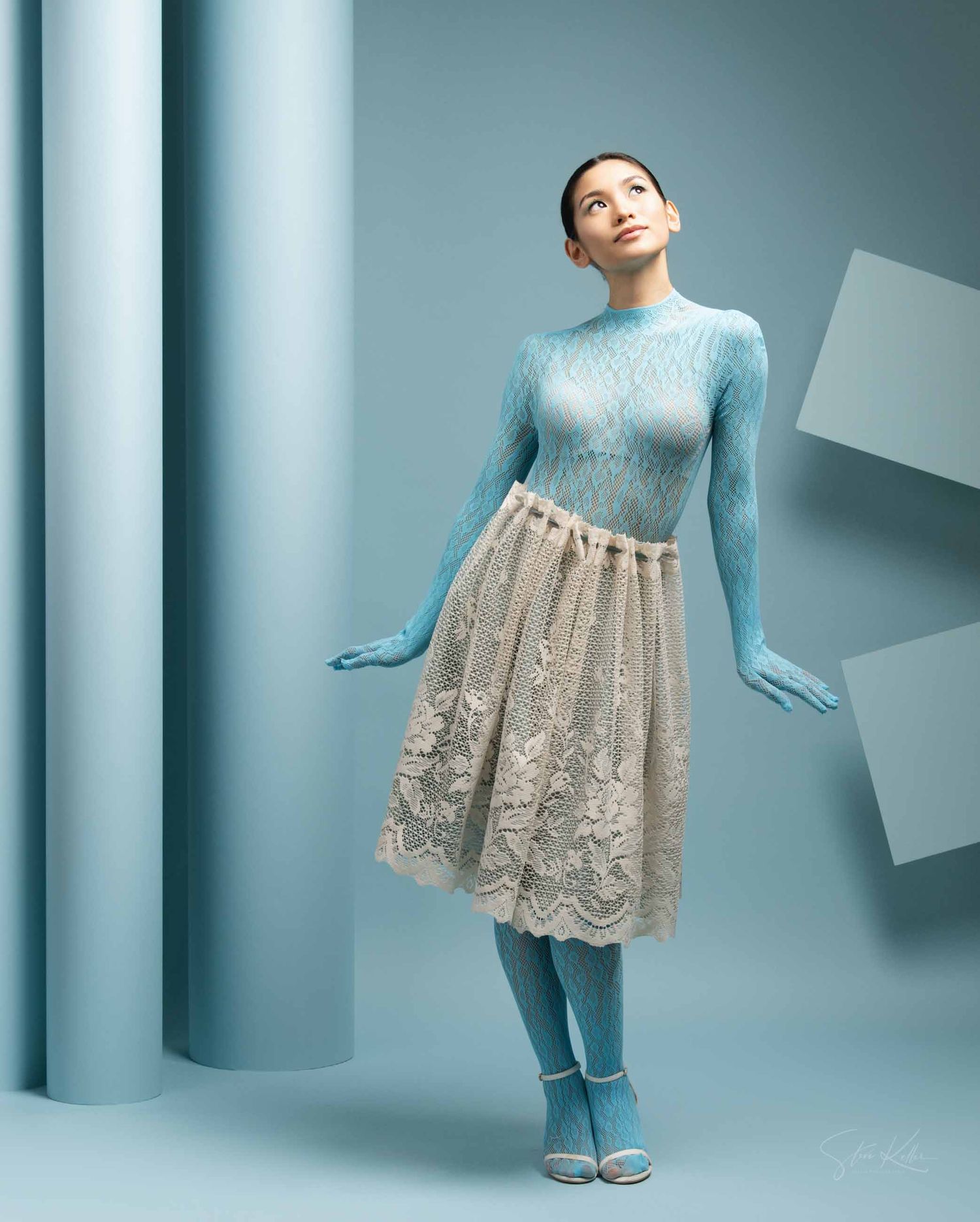 Figure in light blue sweater and lace skirt dress poses gracefully against a blue-tinted studio backdrop.