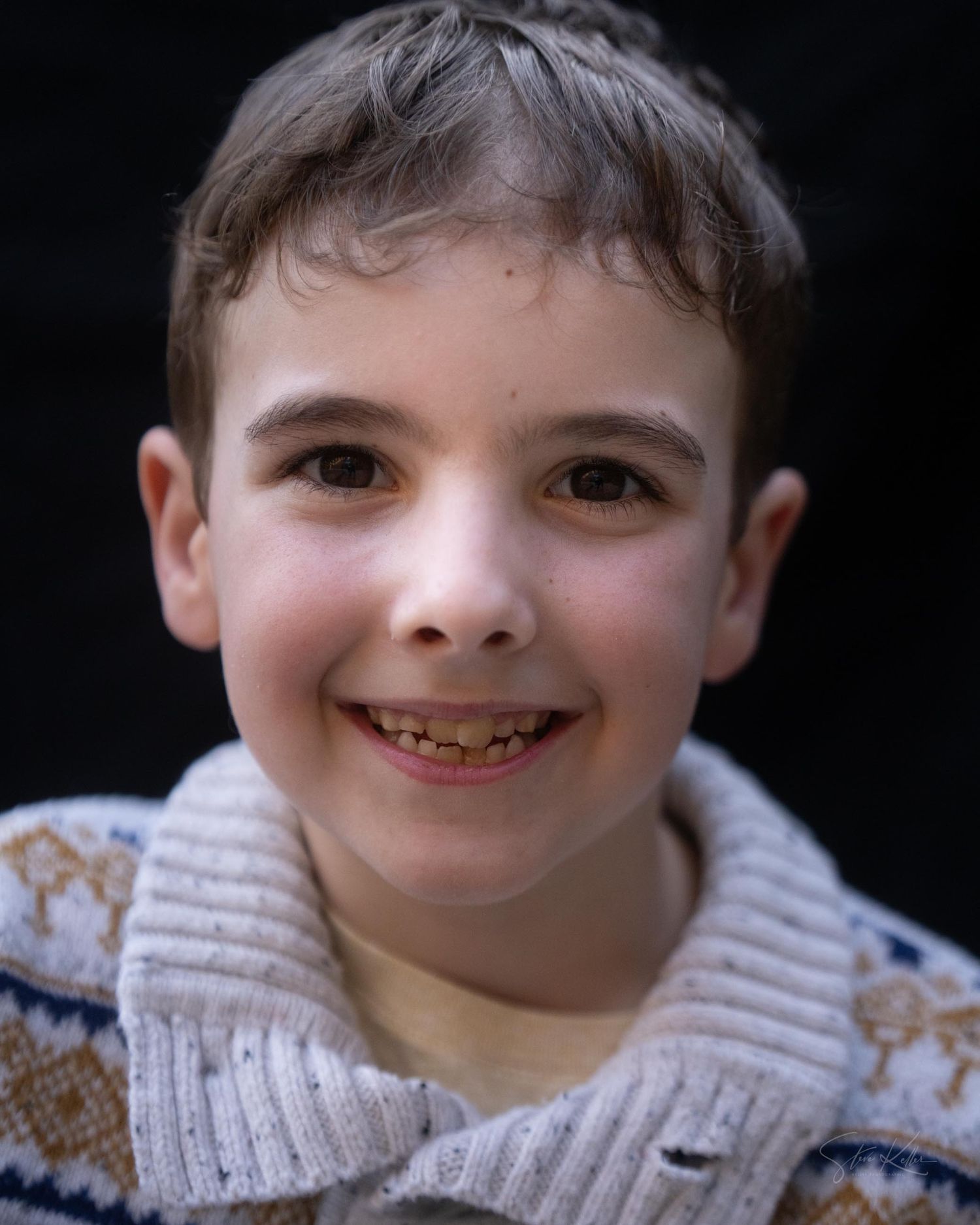 A young child wearing a patterned knit sweater smiles warmly at the camera against a dark background.