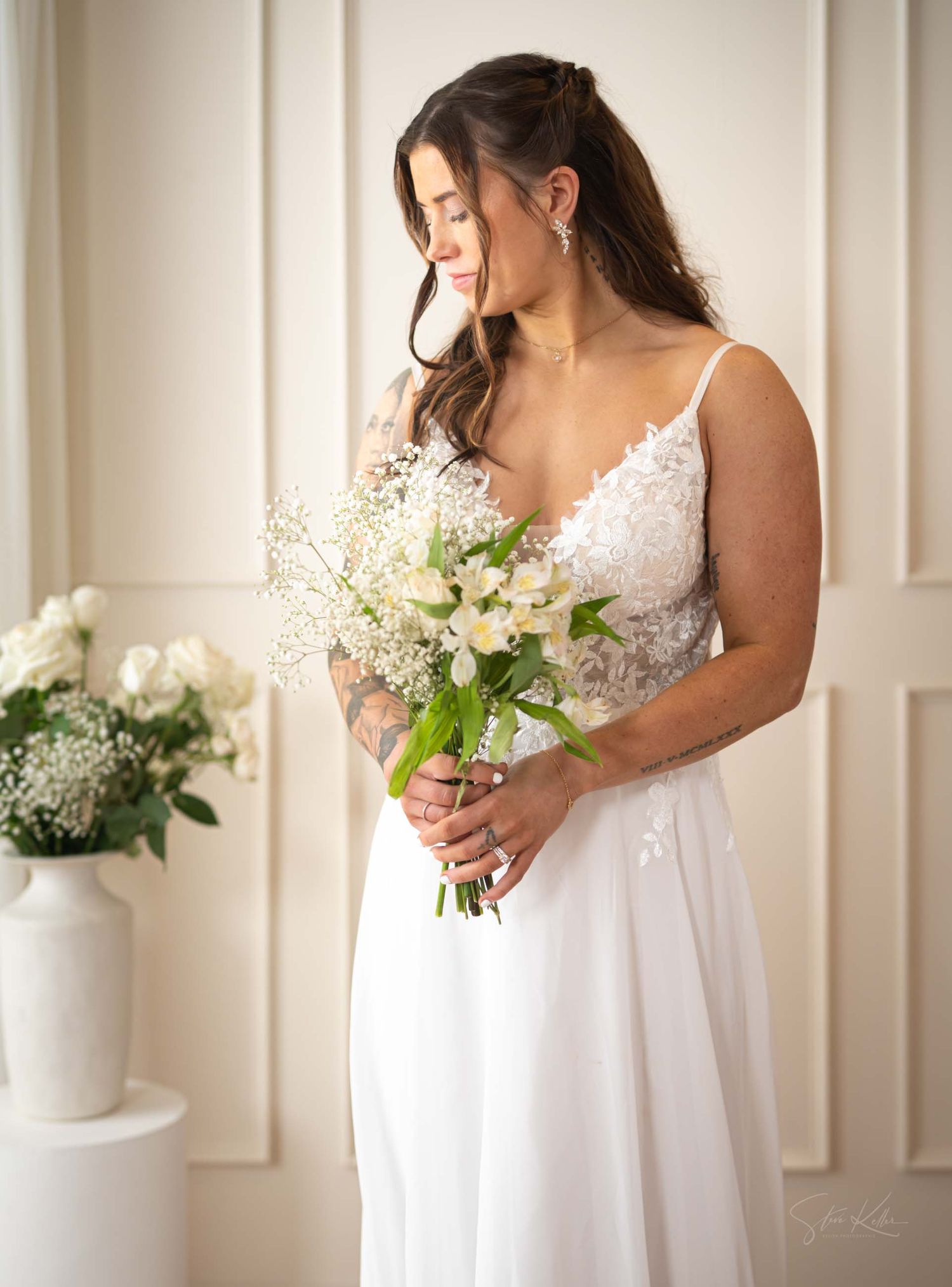 A bride in a white lace wedding dress holds a bouquet of white flowers against a light-colored wall.