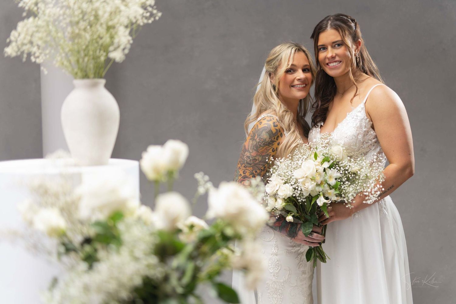 Two people in white wedding attire stand close together with bouquets against a grey background with white floral decor.
