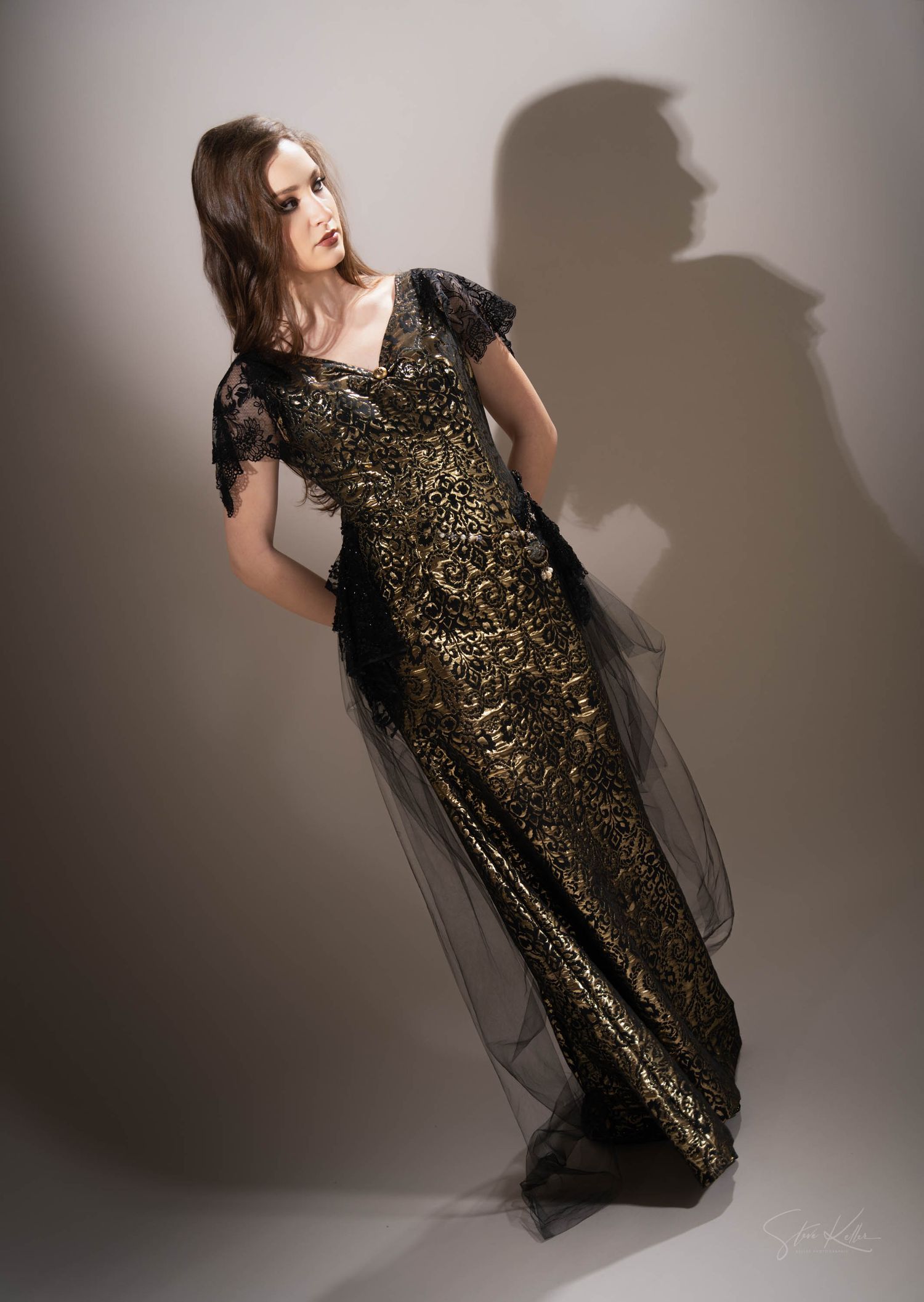 A dramatic fashion photo of a model in a gold and black sequined evening gown with sheer overlay against a shadowy wall.