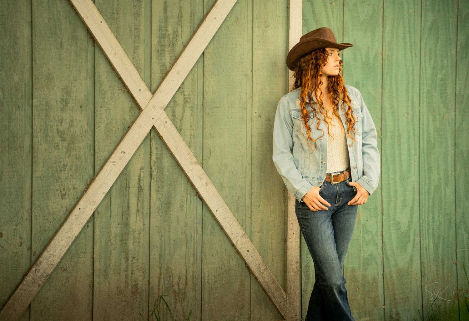 Young woman in cowboy hat, jeans, and denim jacket leaning against a rustic green barn door.