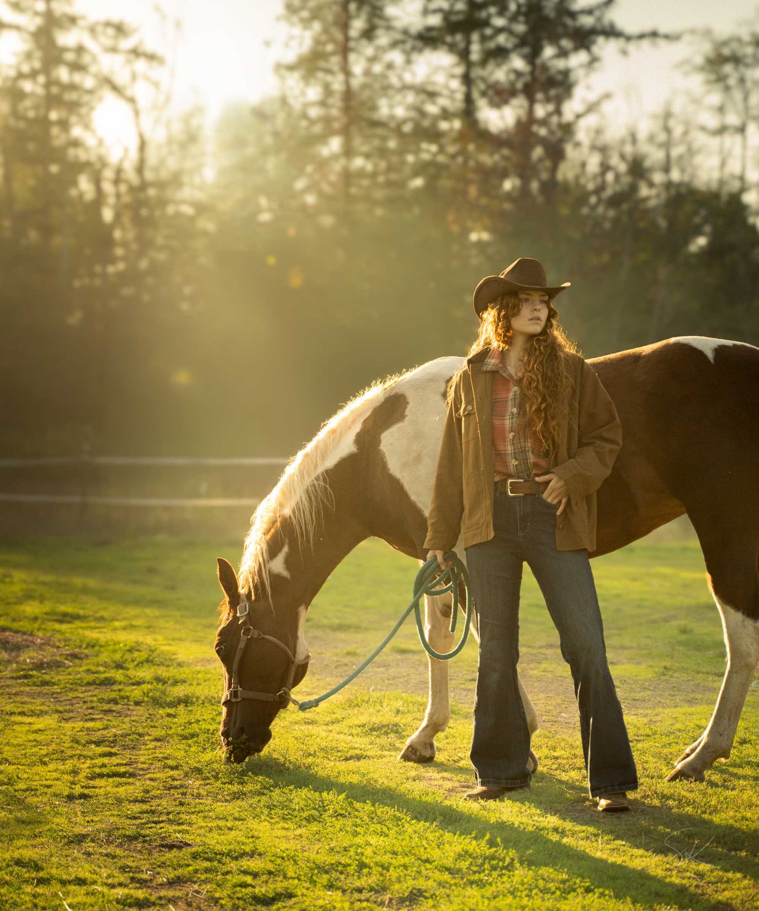 A young woman in a cowboy hat and flare jeans stands with a grazing horse in a sunlit green pasture.