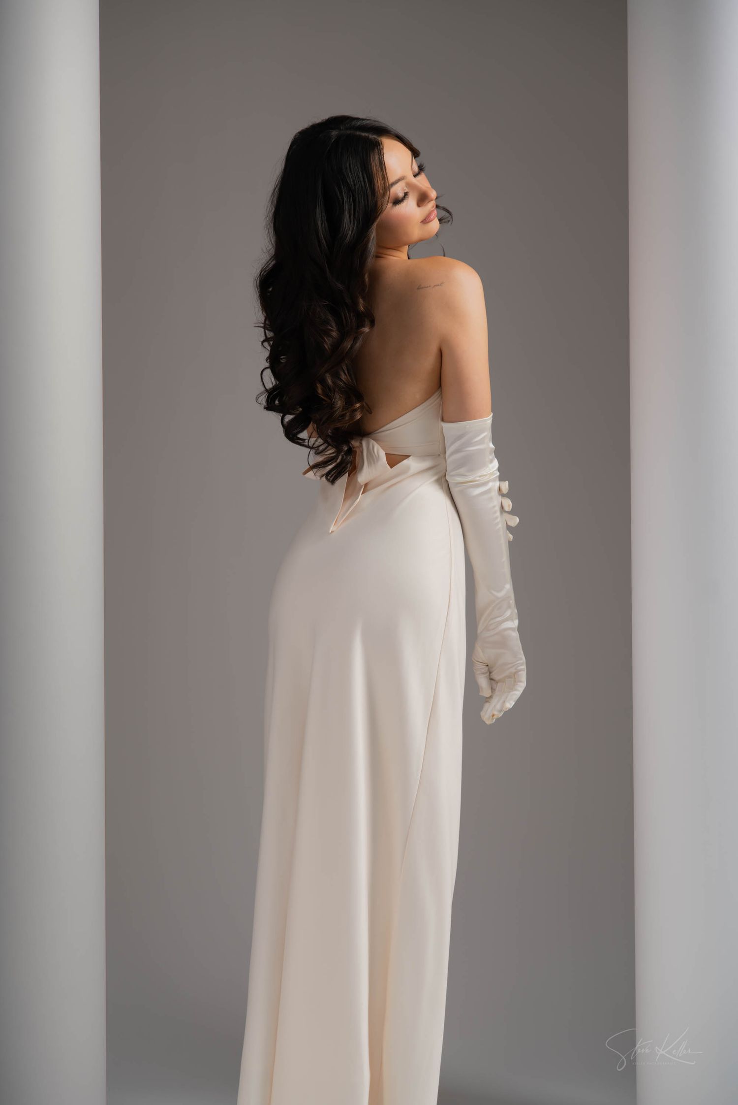 Elegant woman in white evening gown and long gloves poses against grey backdrop between classic columns.