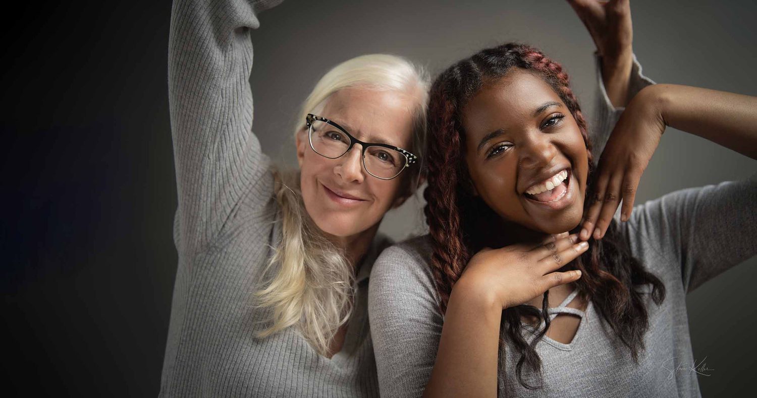Two smiling women posing playfully together with arms raised, one with gray hair and glasses and one with braids, against a dark gray backdr