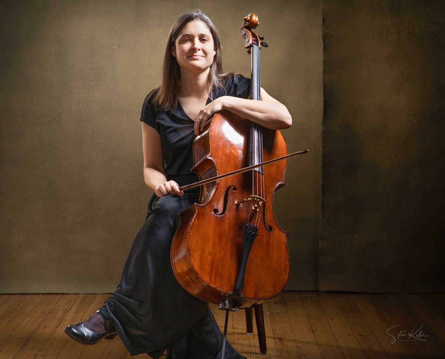 A smiling woman in a black dress poses with a wooden cello in a professional portrait session.