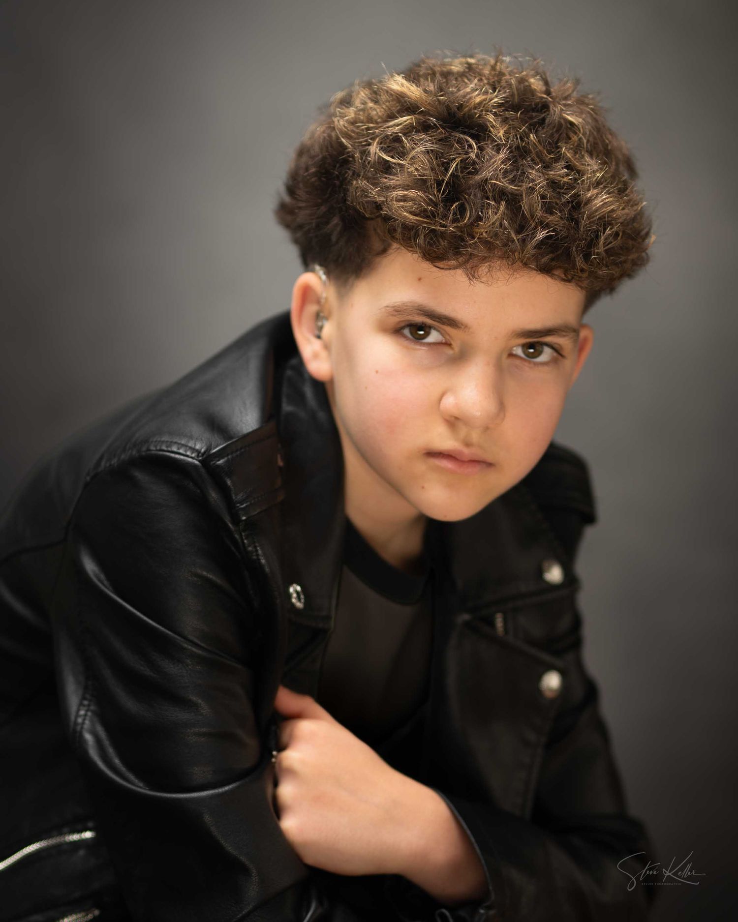 Young boy in black leather jacket with curly hair poses confidently for a professional studio portrait photo.