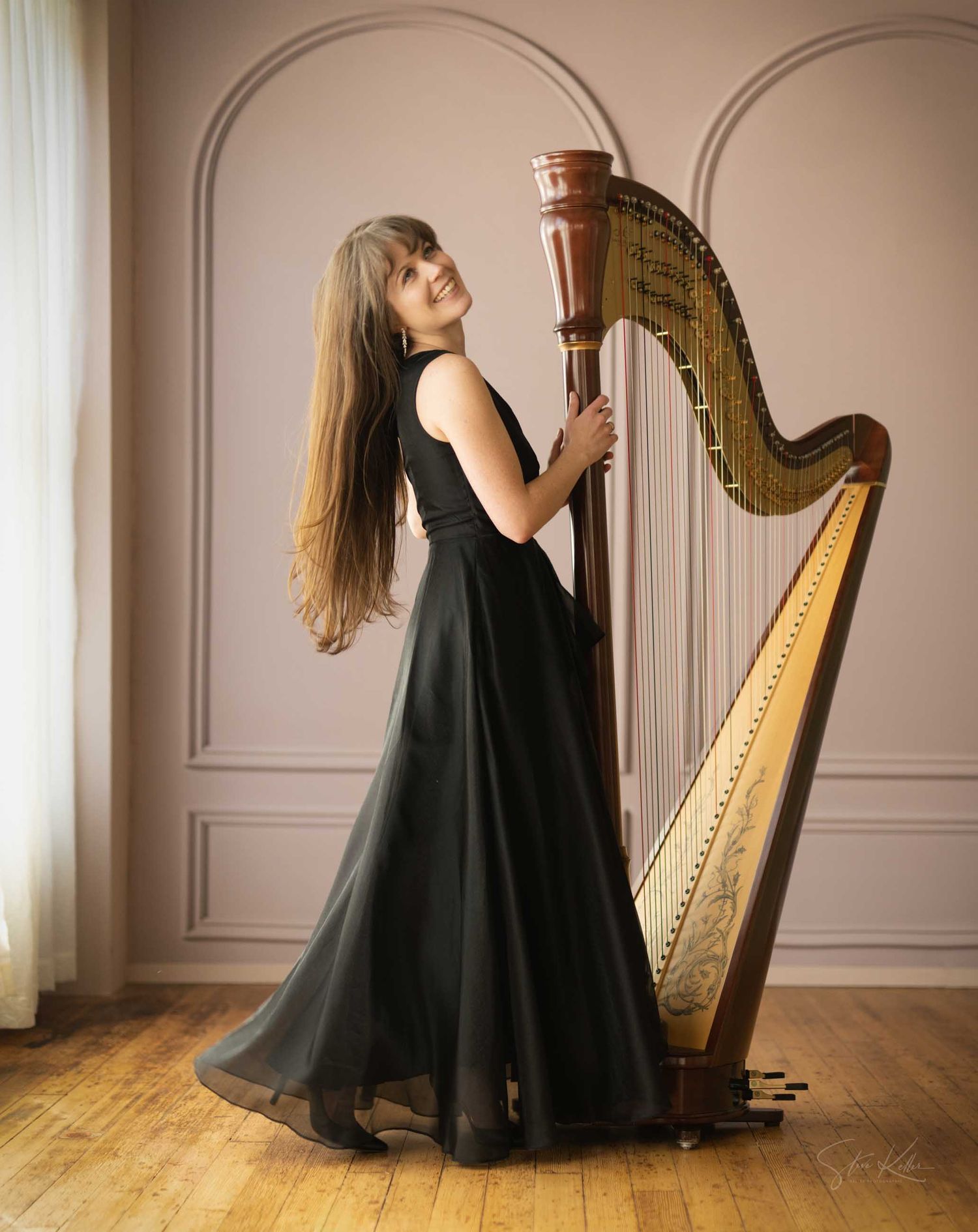 Elegant woman in black gown smiling while posing with a concert harp in a bright, arch-paneled room.
