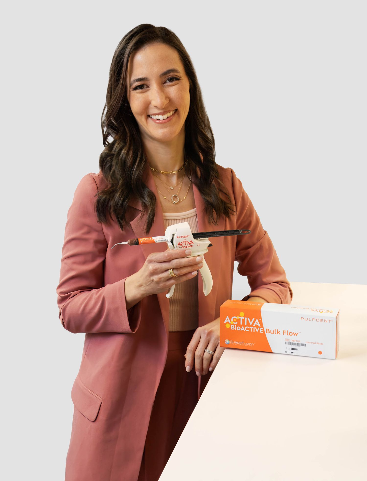 Woman in pink blazer holding ACTIVA BioACTIVE Bulk Flow dental composite syringe beside product box.