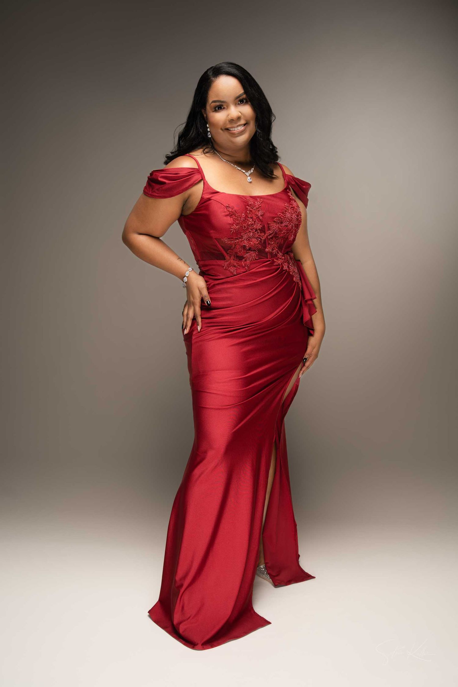 Woman in elegant red satin off-shoulder gown with lace bodice and side slit, posing gracefully in studio.