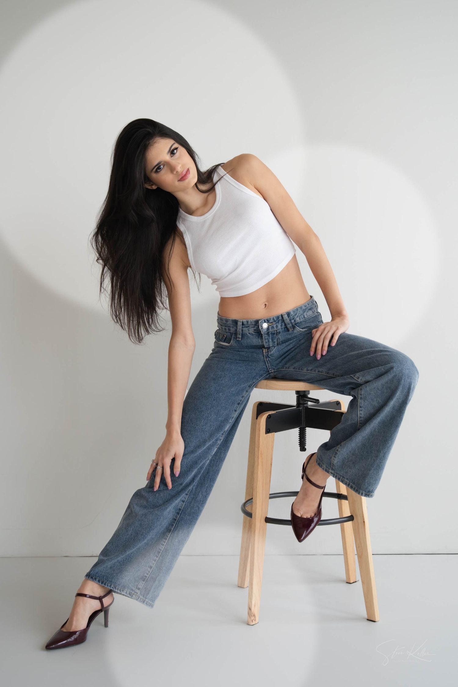 A model in a white crop top and baggy blue jeans poses on a wooden stool against a white background with circular light effects.