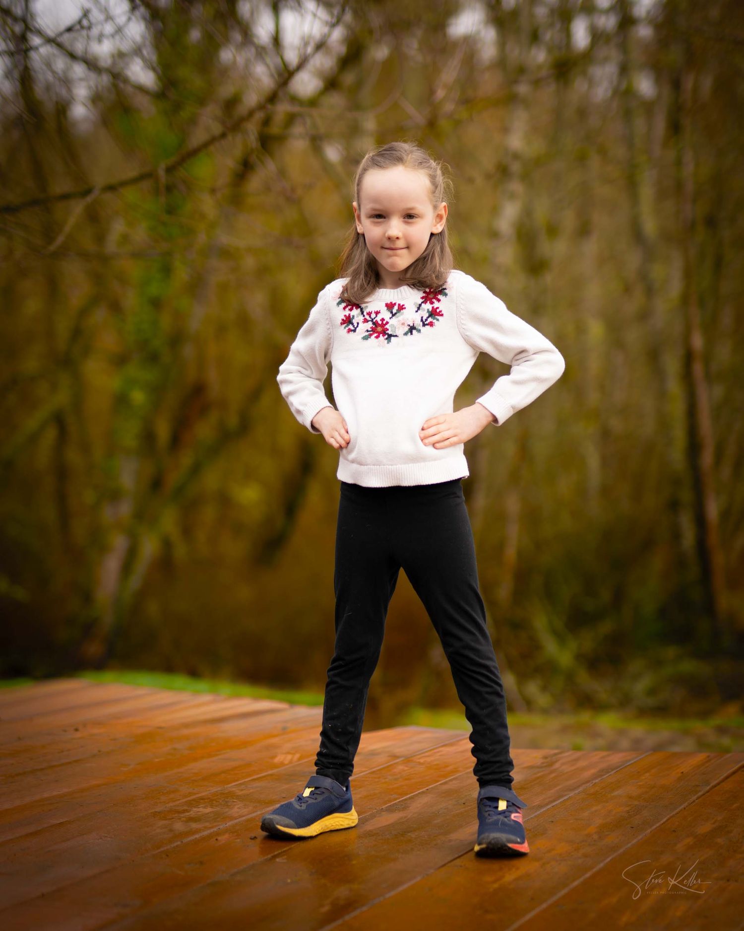 Child in white sweater with floral embroidery and black pants strikes confident pose on wooden deck in forest setting.