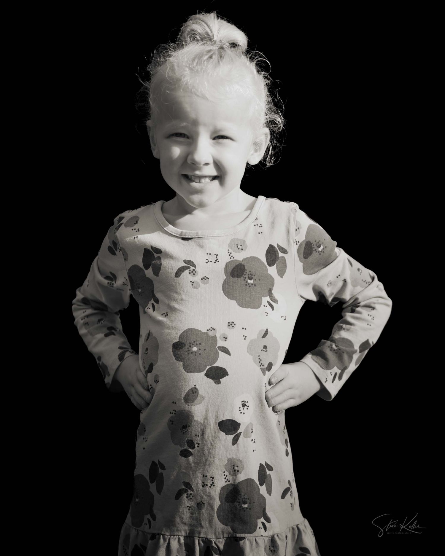A young child wearing a patterned dress smiles and poses against a black background in black and white photography.