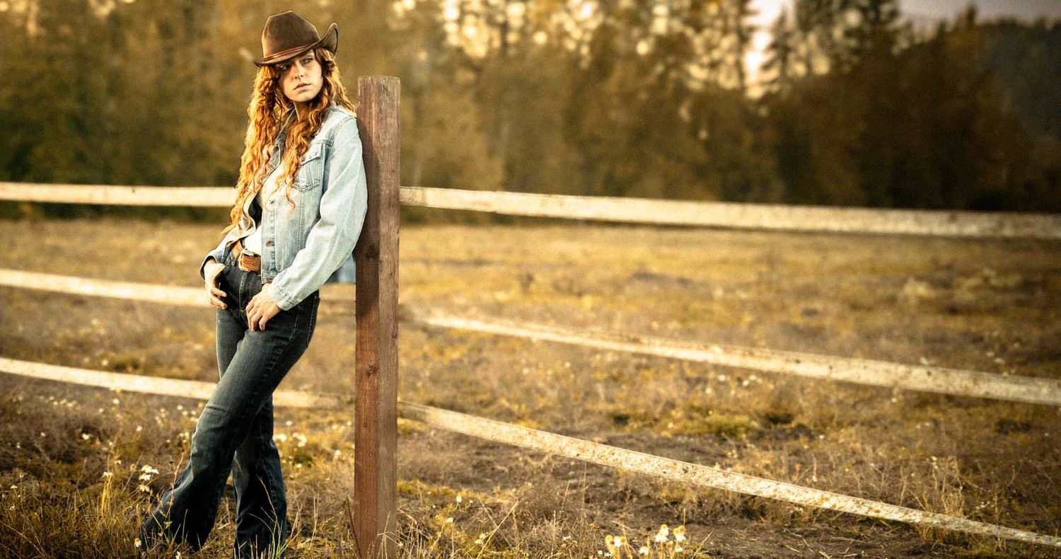 Woman in a cowboy hat and denim jacket leans against a wooden fence post in an autumn field with trees behind.