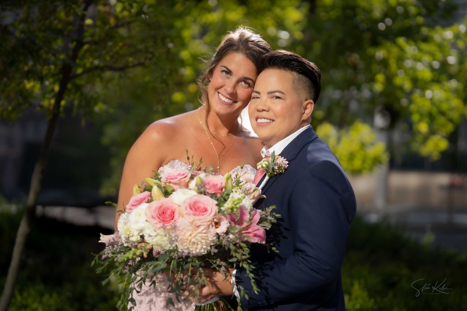 Two brides pose outdoors on their wedding day, one in a white gown holding pink roses, the other in a navy suit.