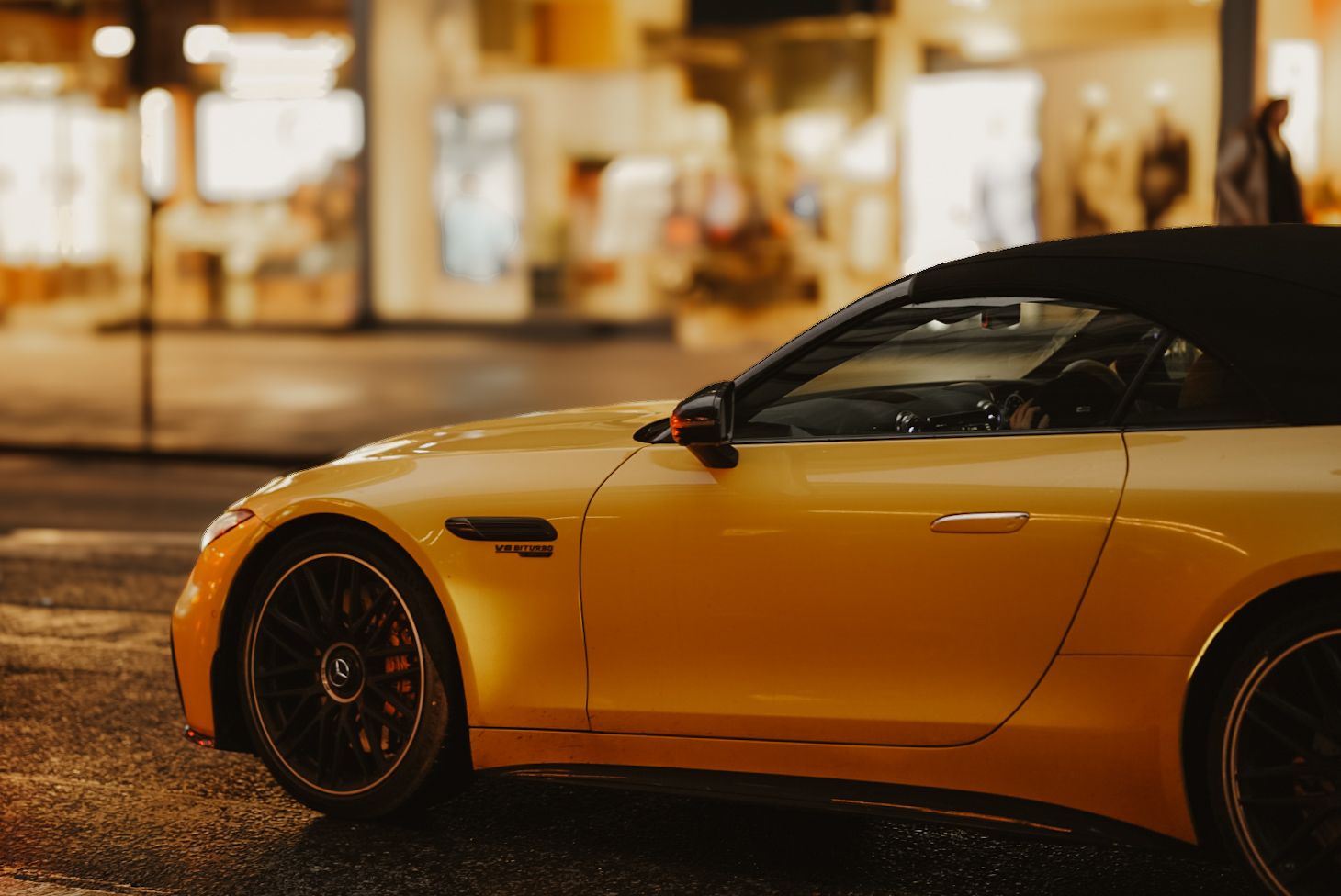 Yellow sports car with black accents photographed at night outside illuminated storefronts.