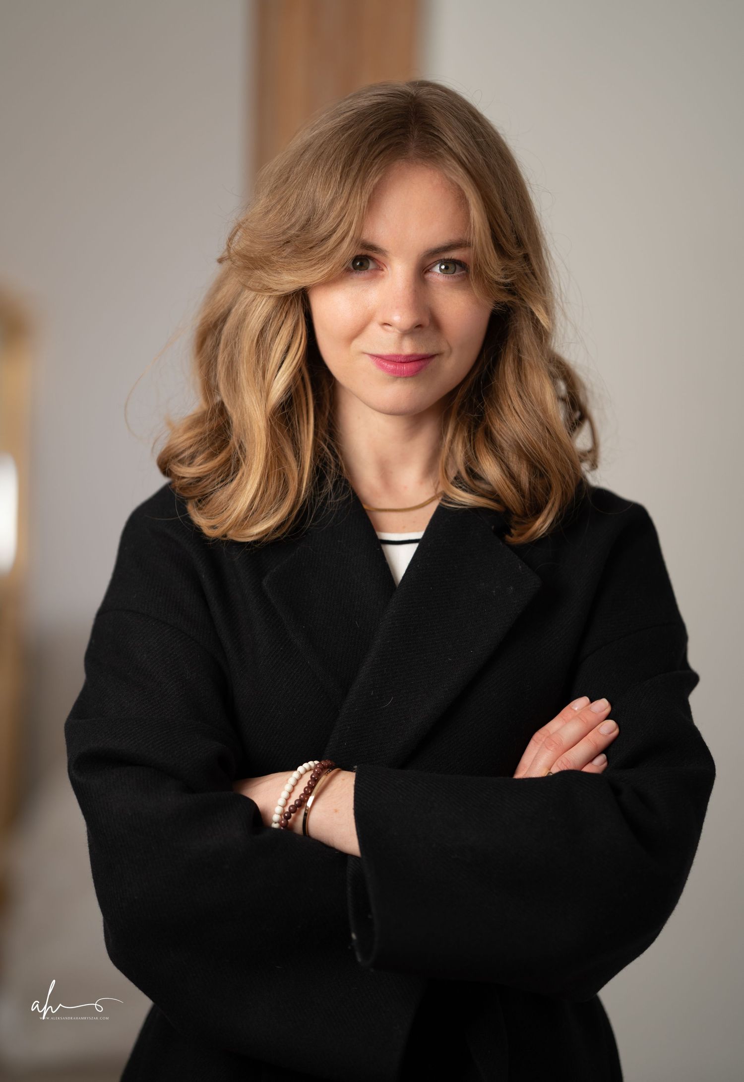 Professional portrait of someone wearing a black coat over white top with styled blonde wavy hair in a warm interior setting.