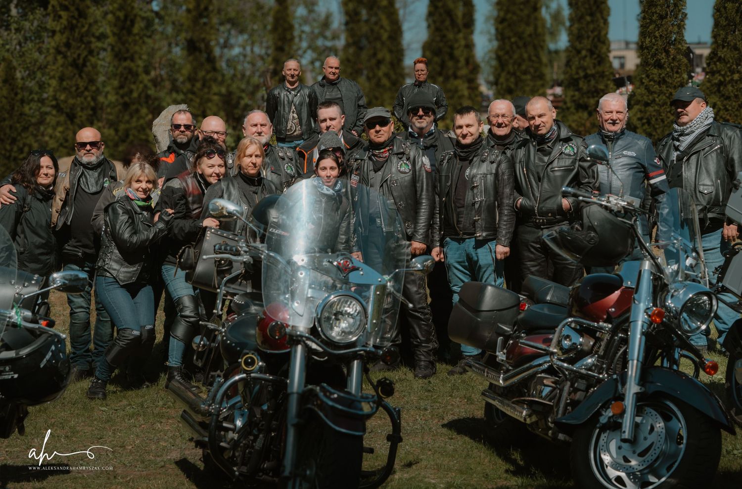 A group of motorcycle riders gathered together at an outdoor event in cloudy weather with bikes parked in rows.