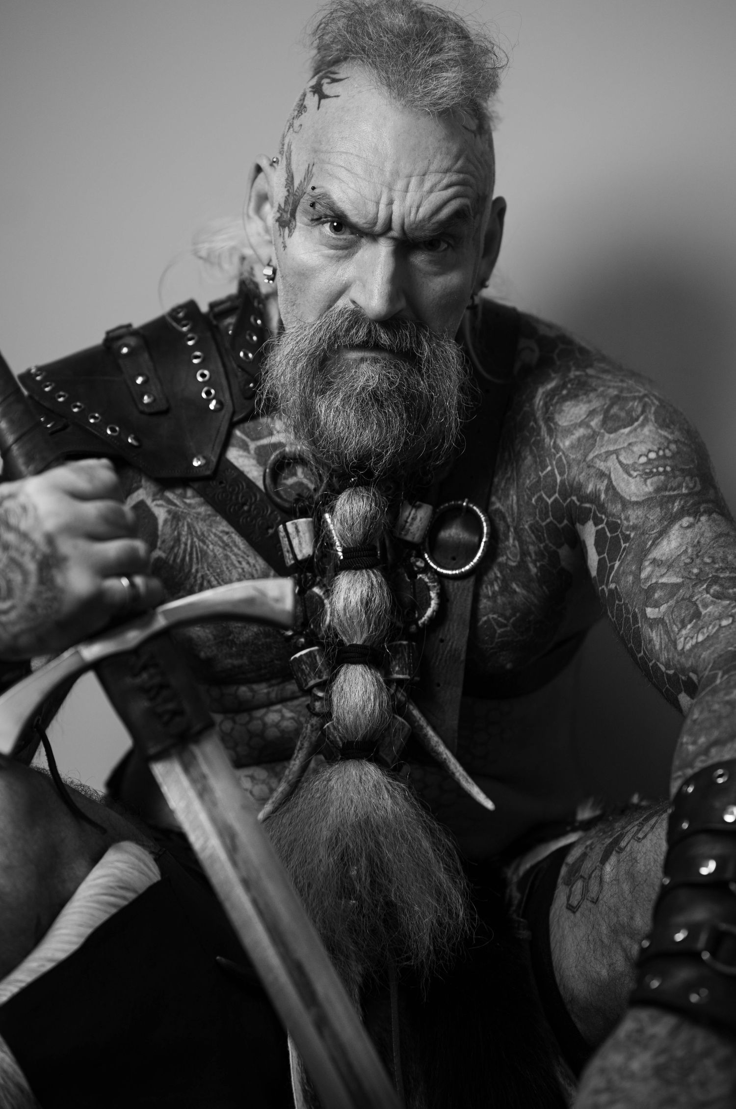 Dark dramatic portrait featuring leather armor, braided beard and medieval-style weaponry.