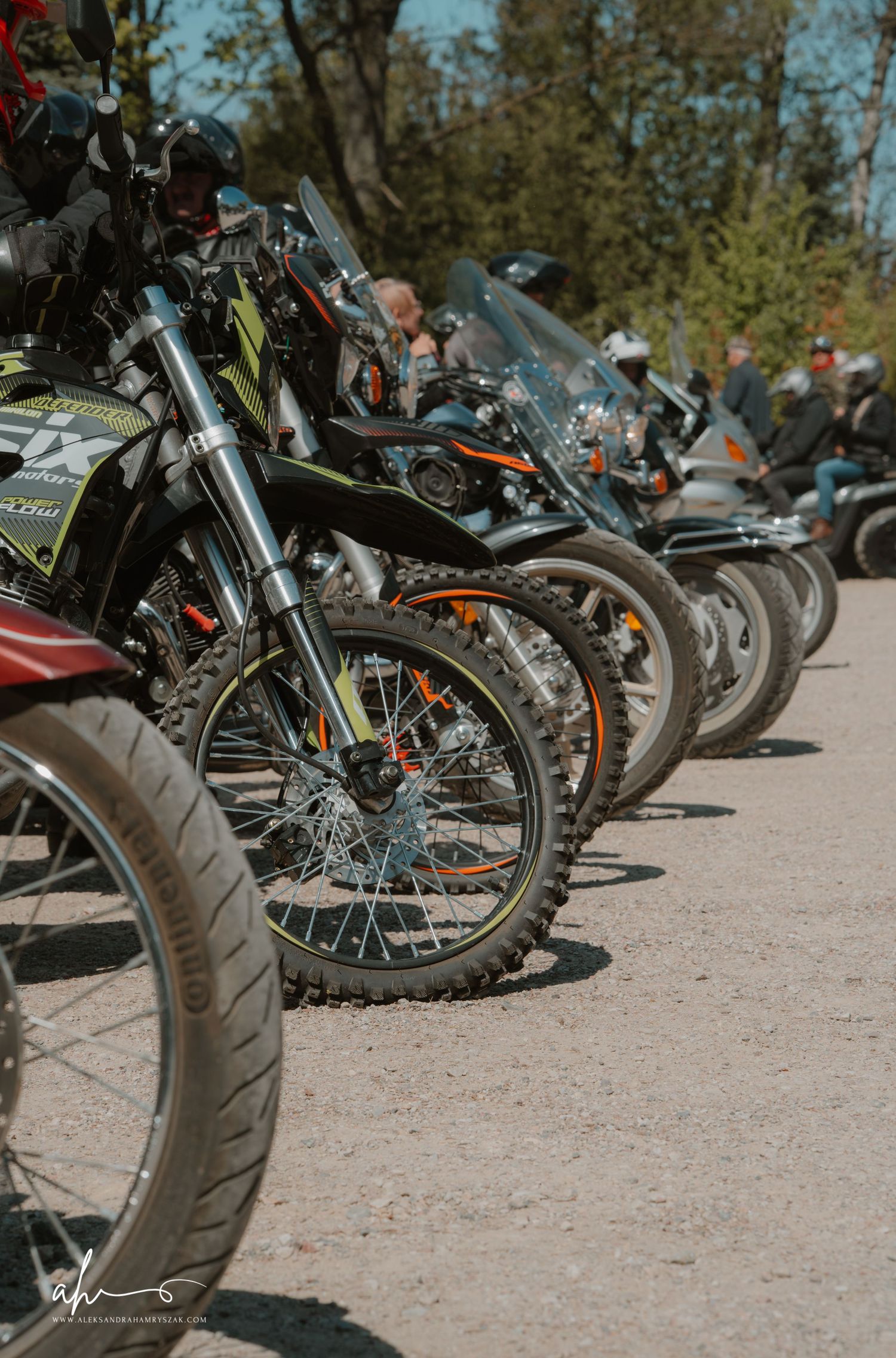Row of parked motorcycles and dirt bikes lined up on dirt ground with trees in the background on a sunny day.