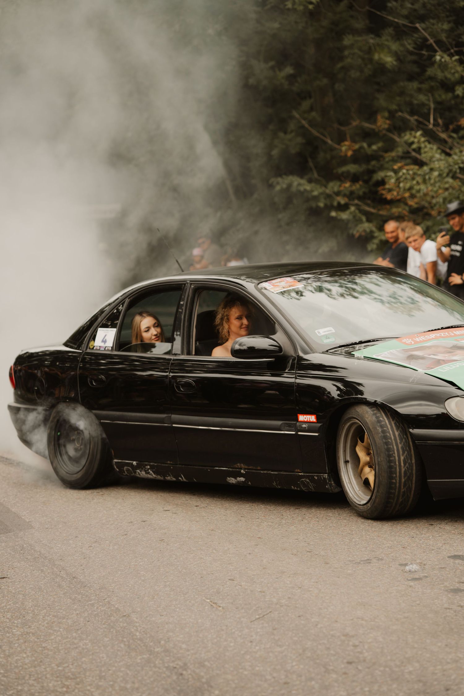 Black sedan performing a smoky burnout on a dirt road with autumn trees in the background.
