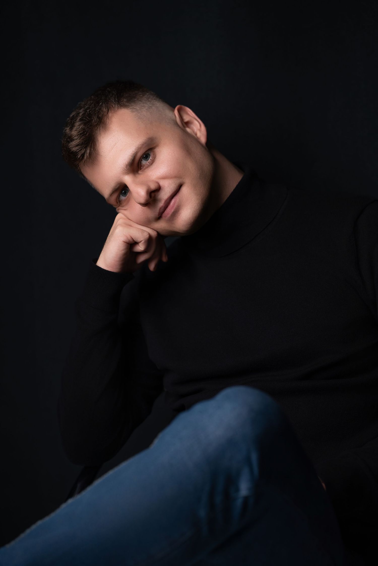 Portrait of someone in a black sweater and jeans against dark background in contemplative pose.