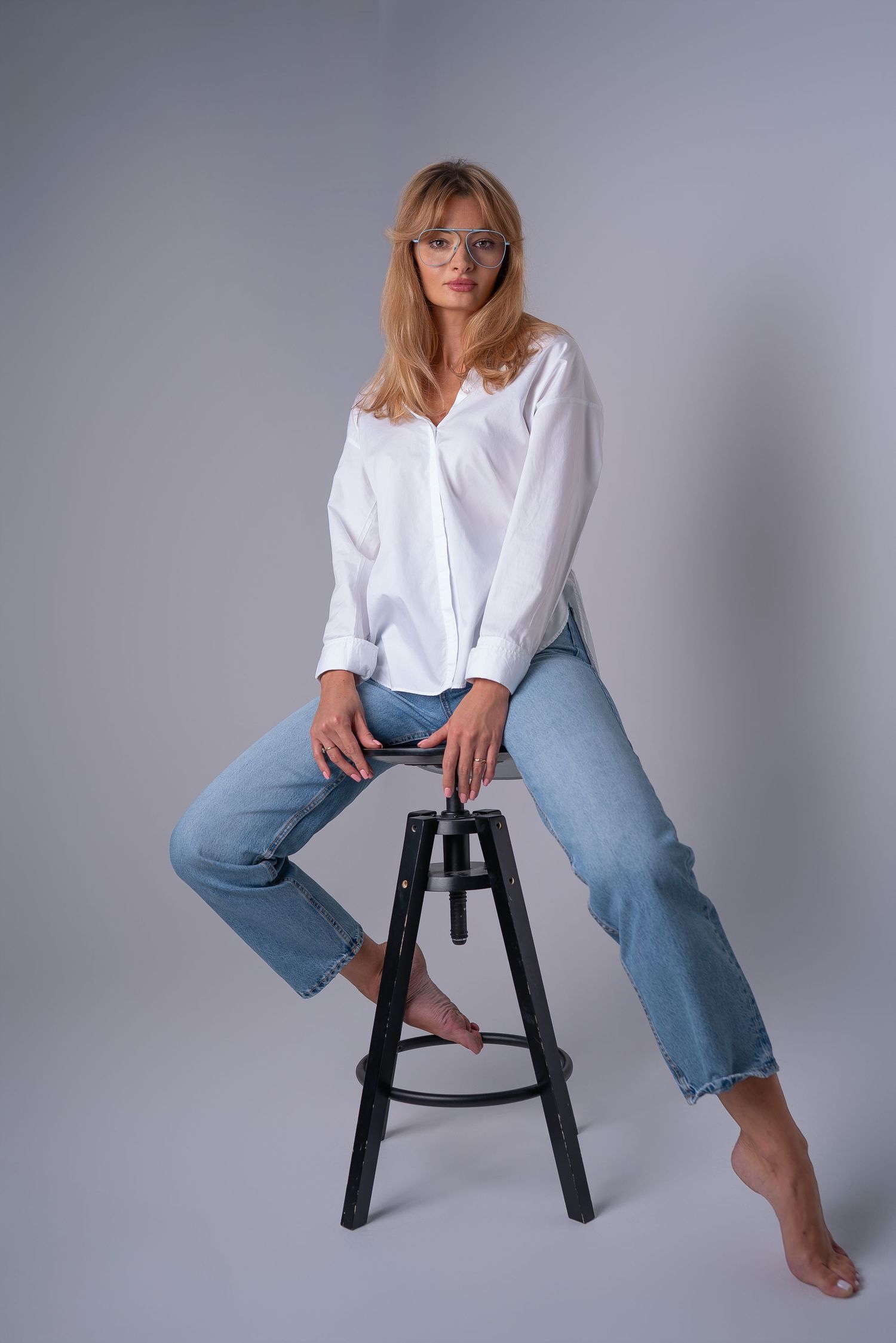 Casual studio portrait with white button-down shirt and relaxed jeans while seated on black stool against gray background.