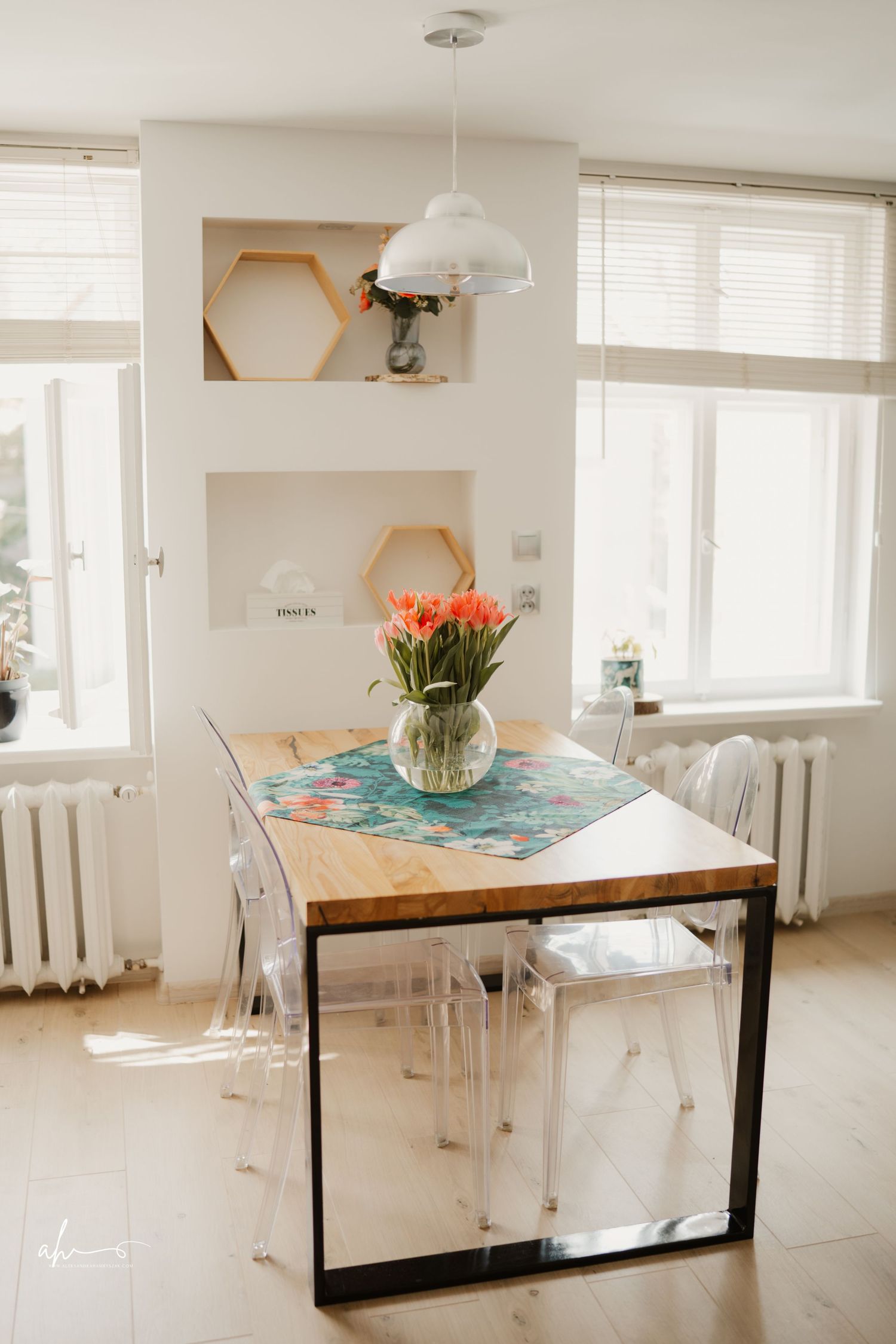 Modern dining space with wood table, clear acrylic chairs, and coral tulips in a bright room with hexagonal wall art.