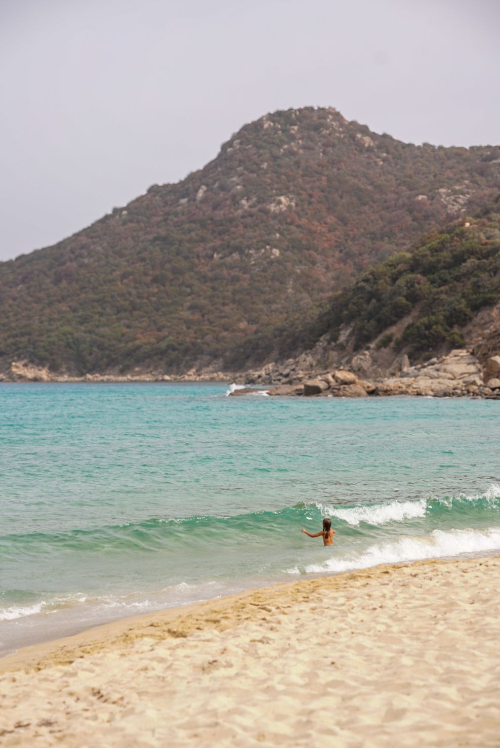 Turquoise waters meet sandy beach with rugged mountain coastline in the background.